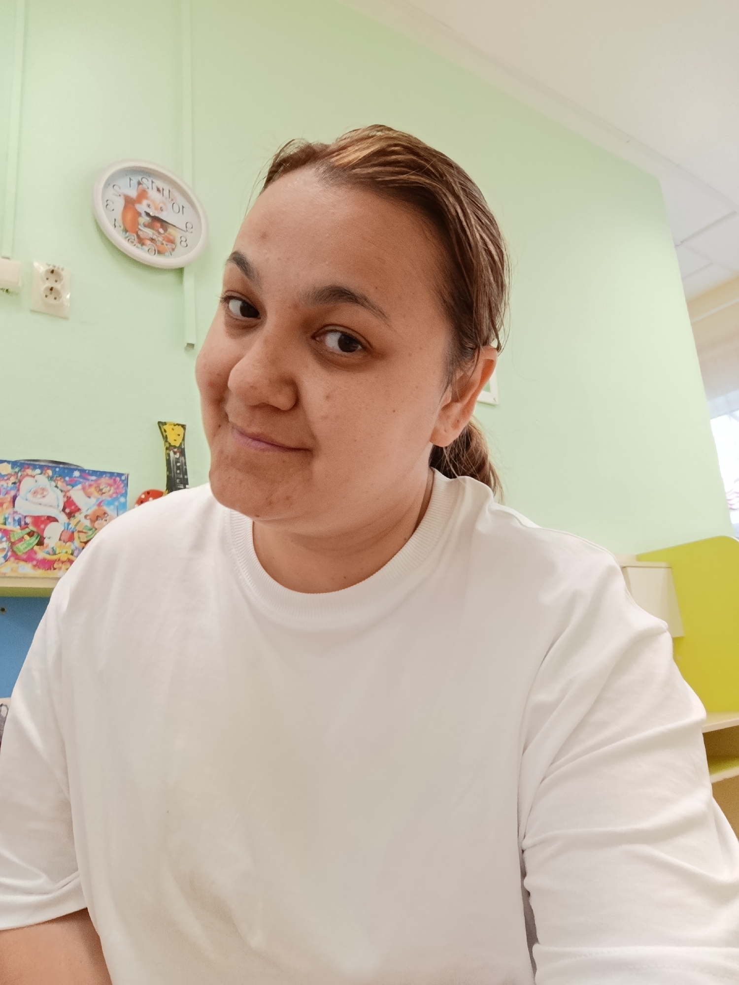 A person with light brown hair, wearing a white t-shirt, is the primary subject of the image. The person is looking at the camera with a slight smile. Behind the person, a light green wall is visible. A clock with an illustration of a fox on its face is mounted on the wall, along with an electrical outlet. A shelf filled with various items, including a Christmas-themed picture and a small yellow object, is also in the background. The location is assumed to be indoors, in the city of Chaikovsky, Russia.