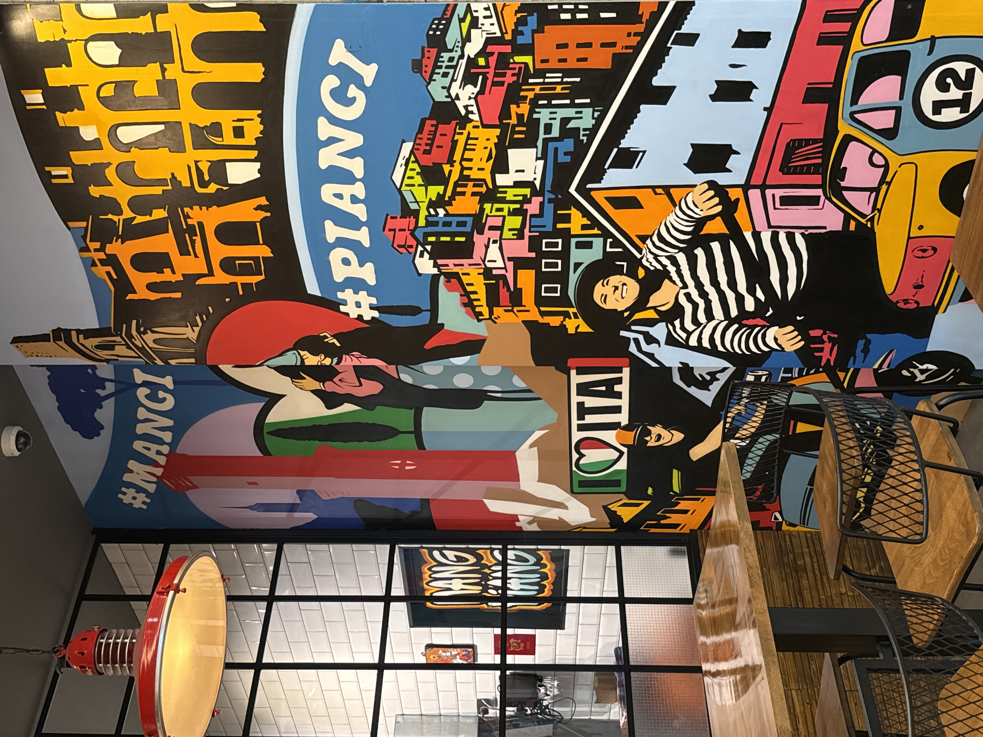 The image shows a section of the interior of a restaurant. A mural on the wall features imagery of Italian landmarks and culture. The Coliseum, colorful buildings, and a person in a striped shirt are depicted. Hashtags #PIANGI and #MANCI, along with "I love ITALIA" are also visible on the mural. In the foreground, there are metal chairs with wooden seats and a wooden table. A red and silver industrial-style lamp hangs from the ceiling. A window with a black frame and white tile walls is visible. The location appears to be a restaurant, likely an Italian restaurant, and is in Sofia, Bulgaria.
