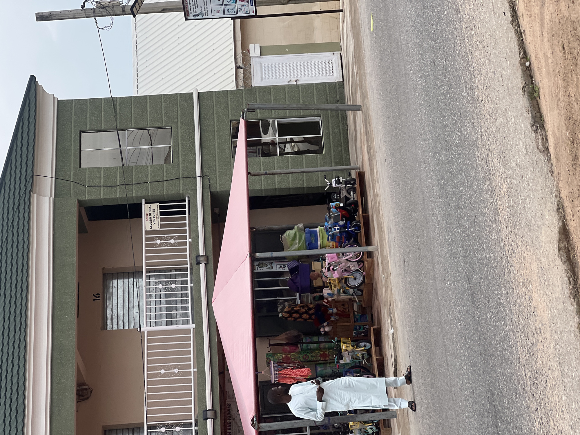 In Jalingo, Nigeria, a man wearing a light blue traditional outfit stands on a paved road in front of a shop. The shop is covered by a pink awning and displays children's bicycles, clothing on racks, and various other items. The building behind the shop is painted green, with a white railing on the second floor and windows with metal shutters. A sign on the second floor reads "16". To the right of the green building, another building with a white corrugated roof is visible. A street light pole with electrical wires is positioned near the right edge of the frame. The scene appears to be a commercial area.
