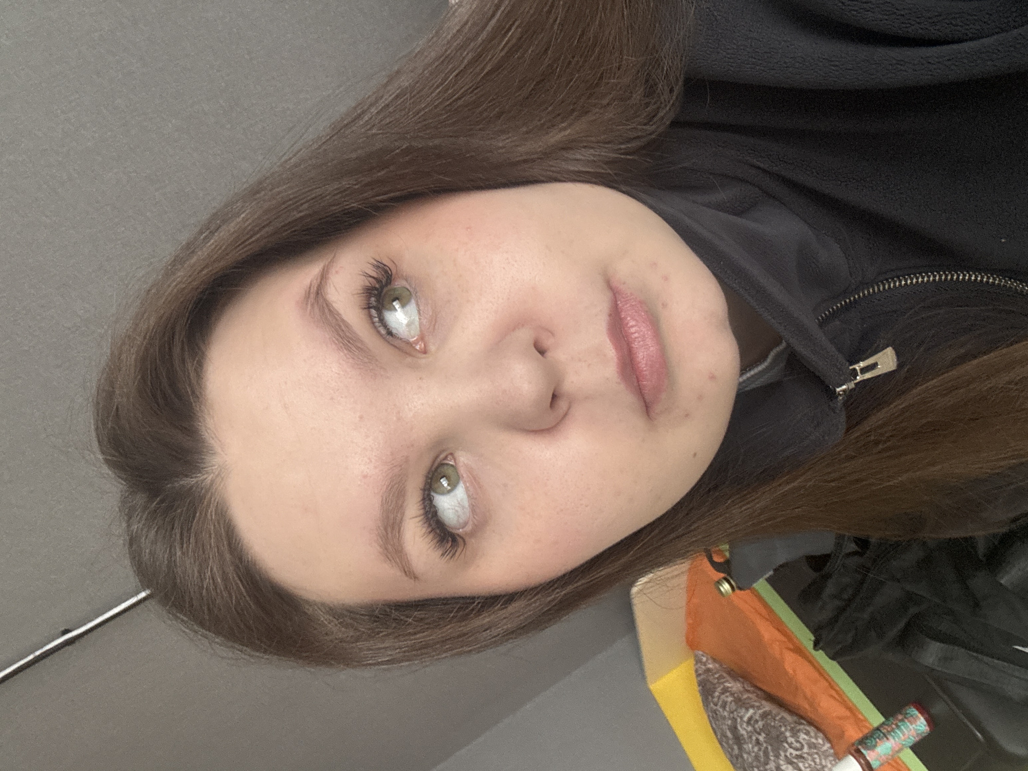 A close-up, head-and-shoulders shot features a young woman with light skin and brown hair. Her eyes are light green, and she has long, dark eyelashes. She is wearing a black zippered fleece jacket. The background includes a grey wall and glimpses of other objects, including a yellow surface and what appears to be clothing. There is no definitive evidence to determine the location as Chaikovsky, Russia.