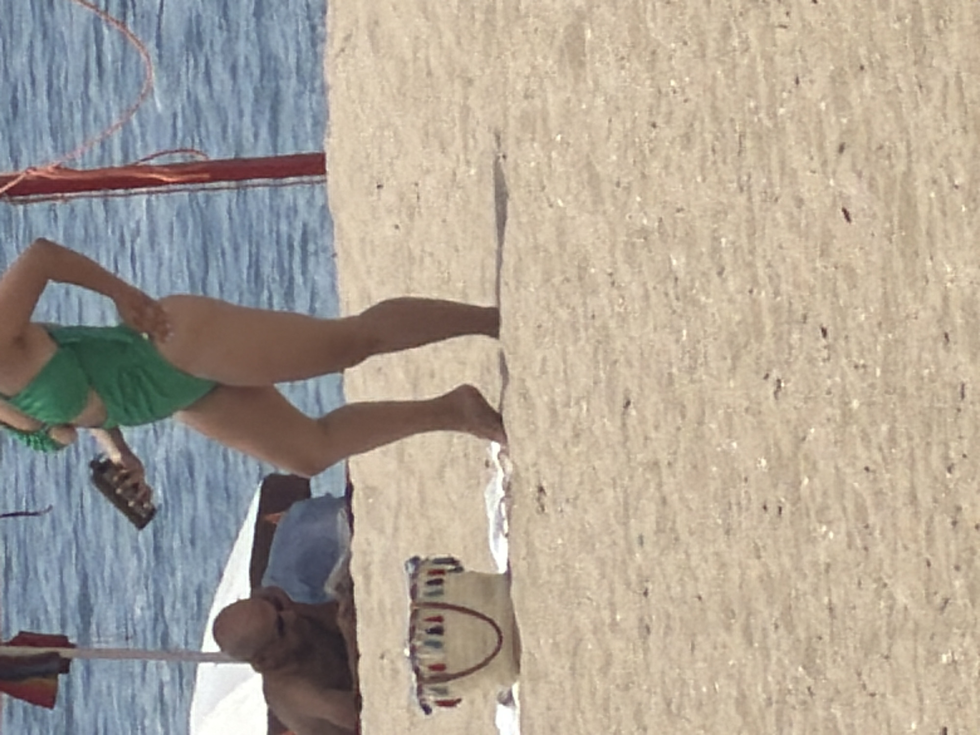 In Espinho, Portugal, a woman in a green swimsuit is partially visible from the waist down on a sandy beach. She is holding a dark-colored object in her right hand. To her left, a man in sunglasses is lying down and appears to be reading. A straw beach bag with colorful tassels is placed on the sand in the foreground. The sea is visible in the background.