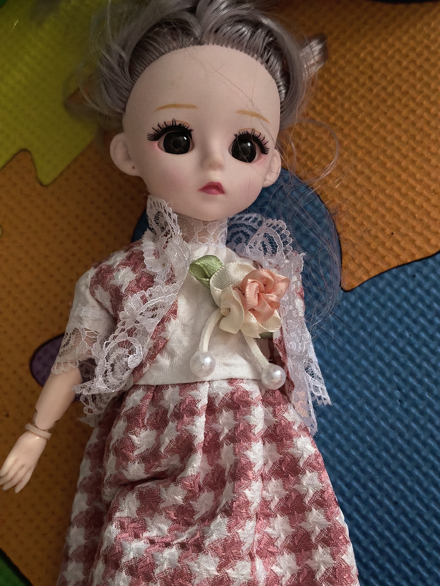 A doll with gray hair and pale skin is lying on a multicolored surface. The doll is wearing a dress with a pink and white houndstooth pattern, a white blouse, and a short jacket with lace trim. A flower with pearl accents adorns the blouse. The background appears to be a patterned play mat with sections of blue, orange, and yellow. No specific location or city is identifiable from the image alone.