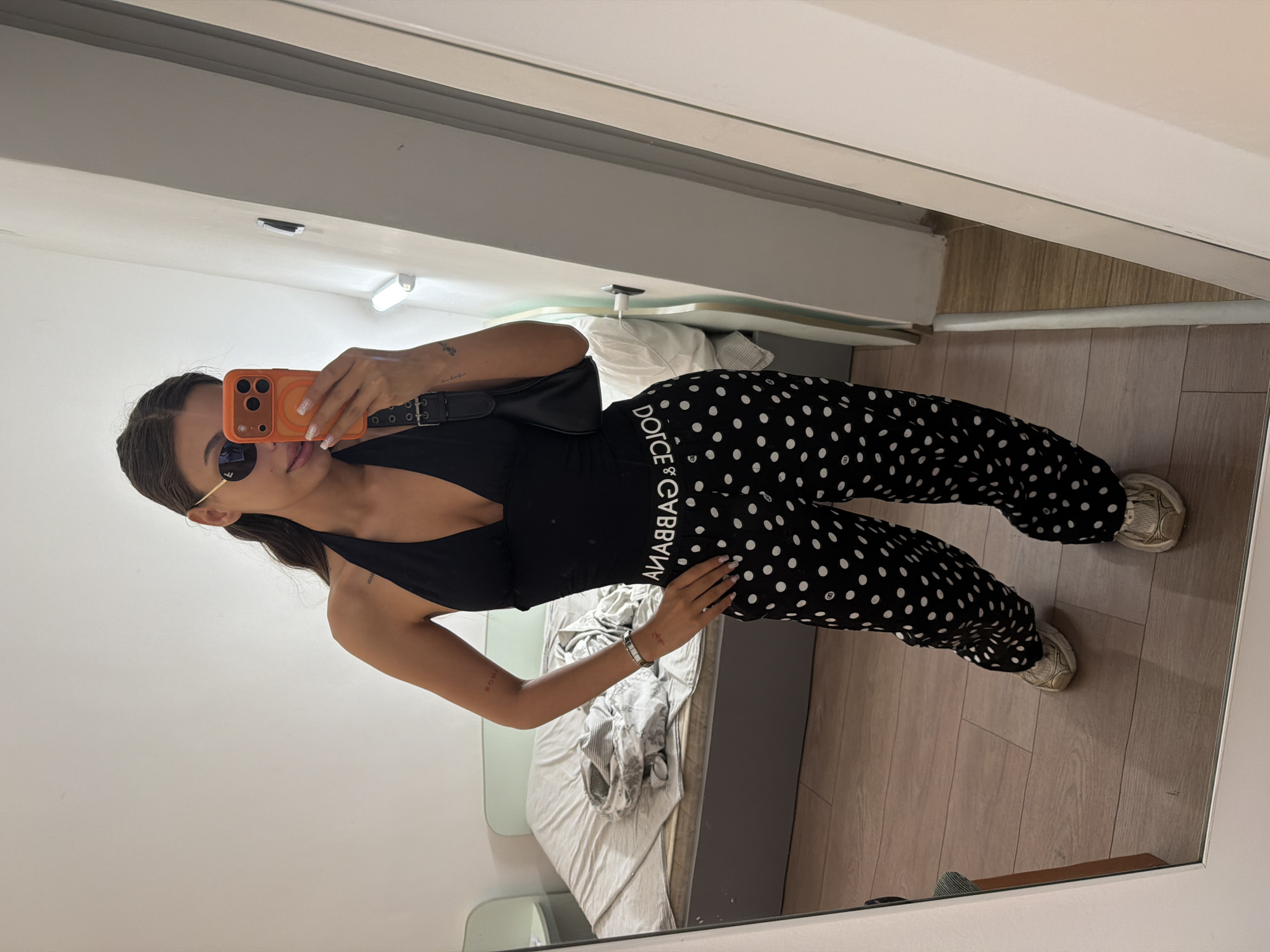 A young woman stands in a bedroom, taking a mirror selfie. She is wearing a black halter top, black and white polka dot pants with "DOLCE & GABBANA" printed on the waistband, and gold sneakers. She has on sunglasses and is holding an orange smartphone in her right hand. A black shoulder bag is slung over her left shoulder. The room has a bed with grey and white bedding, a light blue headboard, and a wooden floor. The scene appears to be set in an apartment in the city of Svishtov, Bulgaria, based on the provided context.
