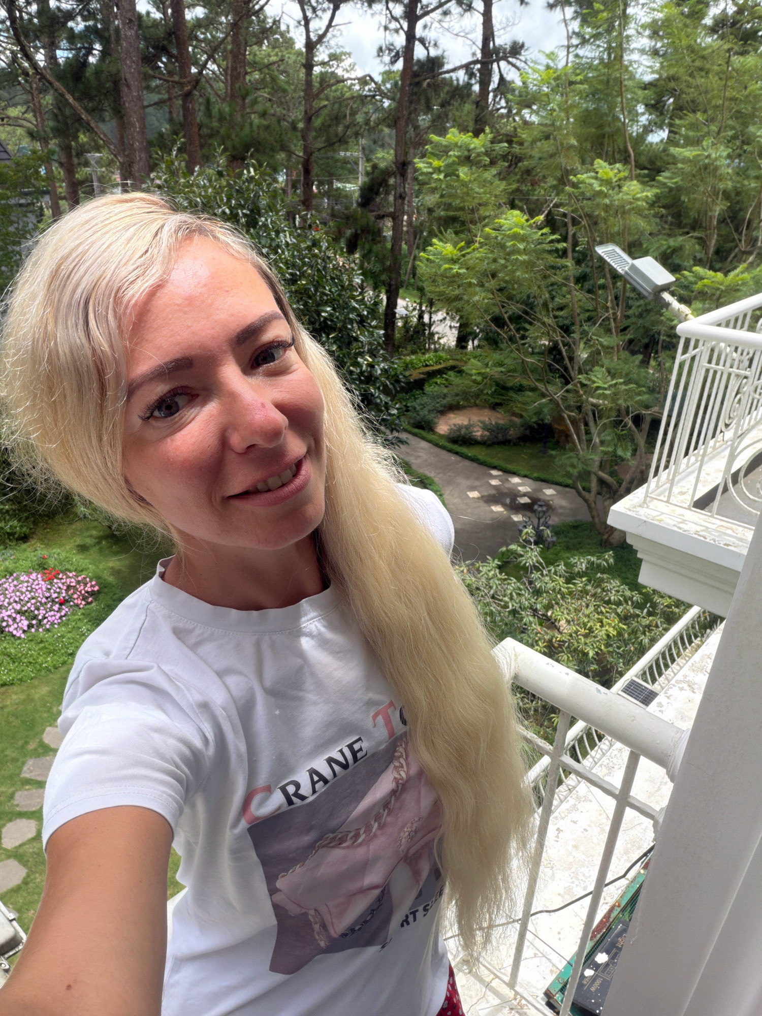 A blonde woman is taking a selfie on a balcony. She is wearing a white t-shirt with a graphic print. The balcony overlooks a garden with trees, a pathway, and a flowerbed. The weather appears to be cloudy. The location is identified as Xuan Thanh, Vietnam.