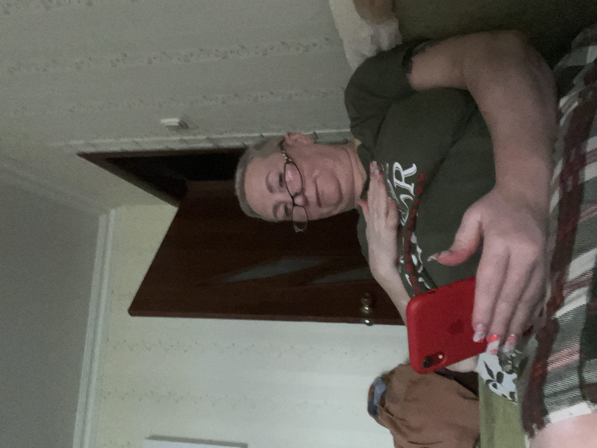 The image depicts an adult female seated indoors, identified as being in Minsk, Belarus. She has short, light-colored hair and wears eyeglasses with dark frames. The subject is wearing an olive green t-shirt with a white capital "R" on the chest, and a plaid-patterned fabric covers her lower body. She holds a red smartphone in her left hand; her fingernails are painted bright pink. Her right hand rests on her chest, and tattoos are partially visible on her right arm. The background features a light-colored wall with a subtle vertical patterned wallpaper, and a dark brown wooden door with a brass-colored round doorknob. A white ceiling with decorative molding is present at the top of the frame. The subject is seated on a green upholstered surface, and a light brown fabric object is visible to her left. The scene is dimly lit.