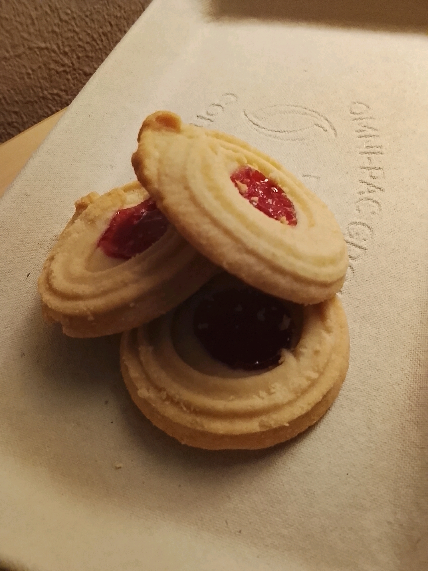 Three shortbread cookies with red jam filling are stacked on a textured light-colored surface, possibly a baking tray or placemat. The cookies are circular with an embossed pattern around the edges and a concave center filled with jam. The lighting suggests an indoor setting, casting shadows on the surface. The background is out of focus and appears to be a darker, textured material. There is no information in the image to determine its location in Malmö, Sweden.