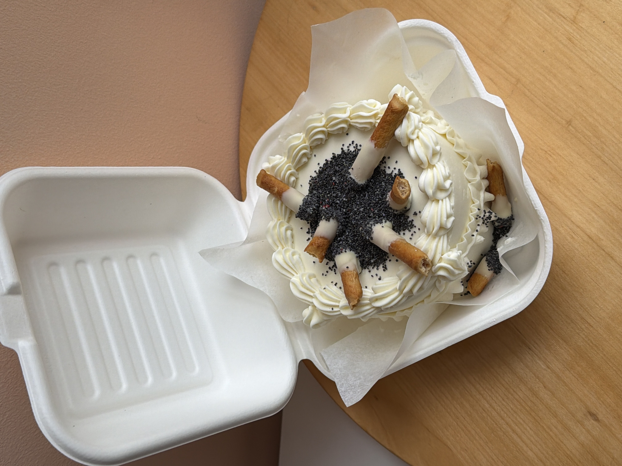 A close-up, high-angle shot shows a cake in a white styrofoam take-out container, placed on a light brown wooden surface. The cake is round and decorated with white frosting swirls around the edge. The center of the cake is covered in black poppy seeds with several cylindrical stick-like decorations, dipped in white frosting, arranged radially around the poppy seeds. The take-out container is partially open, with the lid resting to the left, also in white styrofoam. In the background, there is a tan-colored surface. The setting appears to be indoors. There is no evidence that it is taken in Tel-Aviv, Israel.