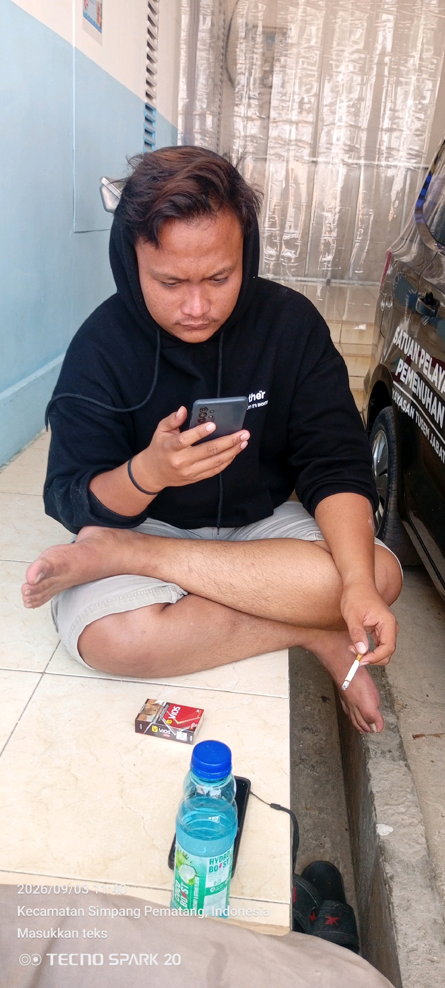 A person wearing a black hoodie and light-colored shorts is sitting cross-legged on a tiled floor, holding a smartphone. A pack of VIOS cigarettes and a bottle of Hydro Coco drink are on the floor to the left of the person. The person is also holding a lit cigarette in their right hand. A black car with text "SATUAN PELAYAN PEMENUHAN YAYASAN..." is partially visible on the right side of the image. The background shows a translucent curtain and a light blue wall. The timestamp "2026/09/03 11:40" and "Kecamatan Simpang Pematang, Indonesia" are overlaid on the bottom left of the image, indicating the location.