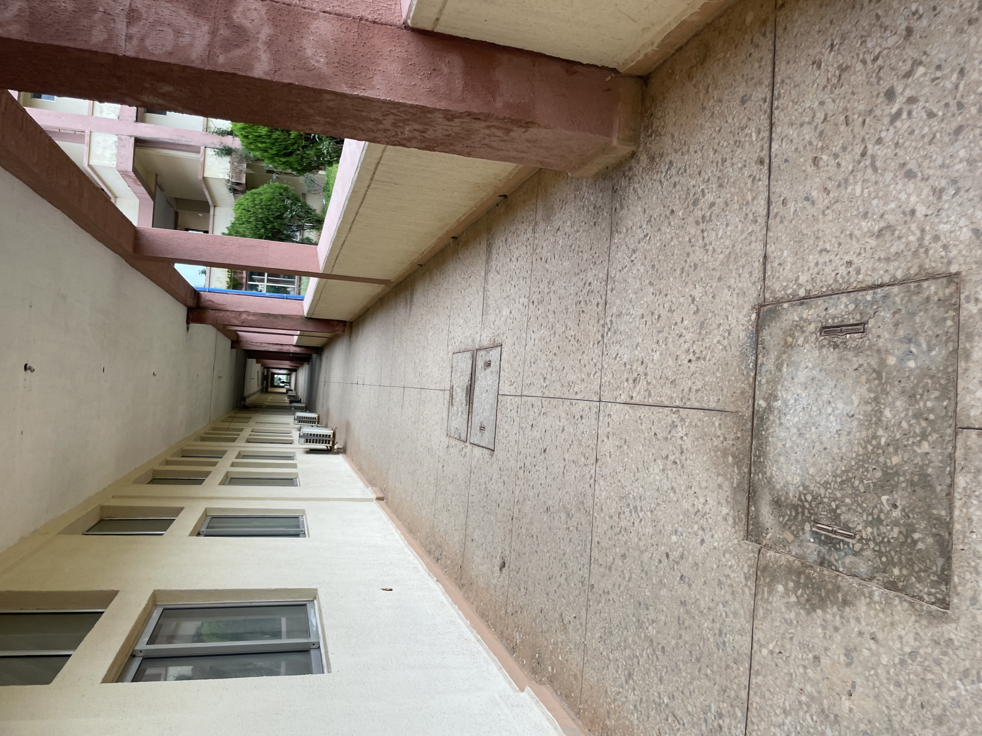 The image shows a long, covered outdoor walkway of a building, likely a school or office complex, in Gwong, Nigeria. The walkway is paved with terrazzo tiles and has a white plastered wall with multiple windows on the left. On the right side, there are structural columns painted in a reddish-brown color supporting the roof. Green bushes and parts of another building with balconies and windows are visible beyond the walkway. Several air conditioning units are placed along the base of the wall. The ground is marked with lines suggesting tiling or expansion joints, and there are at least two square metal utility covers embedded in the pavement. The walkway recedes into the distance, creating a sense of perspective. The lighting suggests it is daytime, possibly overcast. There are no people visible in the image.