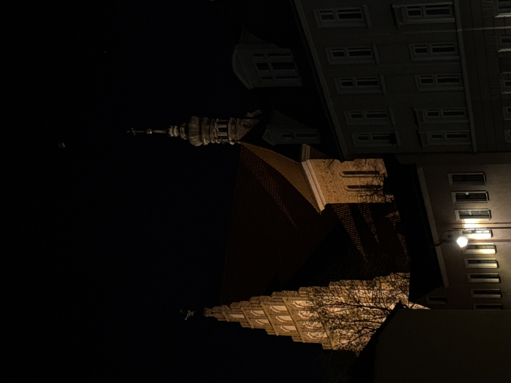 Captured at night in Bydgoszcz, Poland, the image showcases a building with numerous windows and a tall steeple, accompanied by a structure featuring a tiered, decorative roofline. Bare tree branches are visible near the lower right section of the frame. A street light illuminates the lower right building facade.