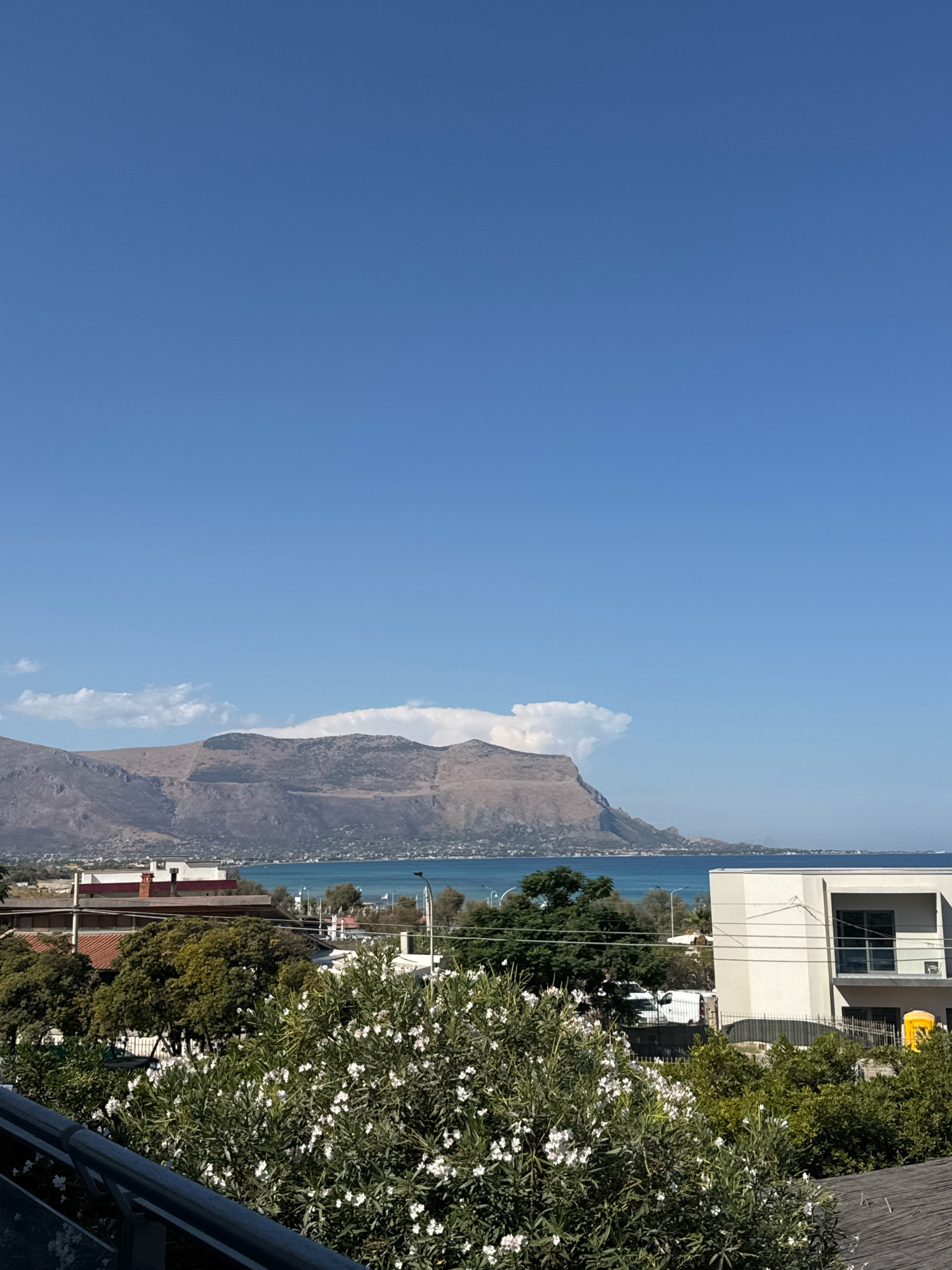 The image depicts a scenic view of the coastline near Capaci, Italy. In the foreground, lush green foliage with white flowers is visible, partially obscuring a railing. Beyond the vegetation, modern buildings with clean lines and balconies can be seen, along with some older structures. A vibrant blue sea stretches towards a distant, rugged mountain range that dominates the horizon. A few white clouds are scattered across the clear blue sky. The overall impression is one of a bright, sunny day in a coastal Italian town, with nature and architecture harmoniously coexisting.