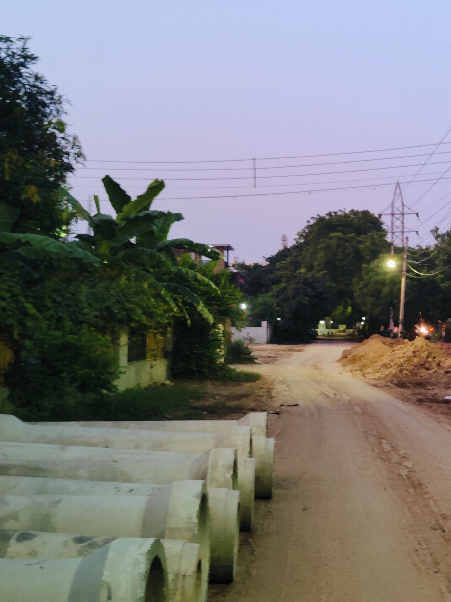 The image depicts a street scene at dusk in Gurgaon, India. In the foreground, a row of large concrete pipes are stacked, indicating potential construction or infrastructure work. The road itself is unpaved and dusty, suggesting it might be a less developed area or a construction site. Lush green vegetation, including banana trees, borders the left side of the street, partially obscuring a small building. In the background, tall trees form a dense canopy, and a tall electricity pylon with overhead wires stands prominently, a common sight in urban and semi-urban Indian landscapes. A street light is illuminated, casting a warm glow on the scene, and piles of dirt or construction debris are visible on the right side of the road. The sky is a soft, pale purple, typical of twilight. There are no people visible in the image.