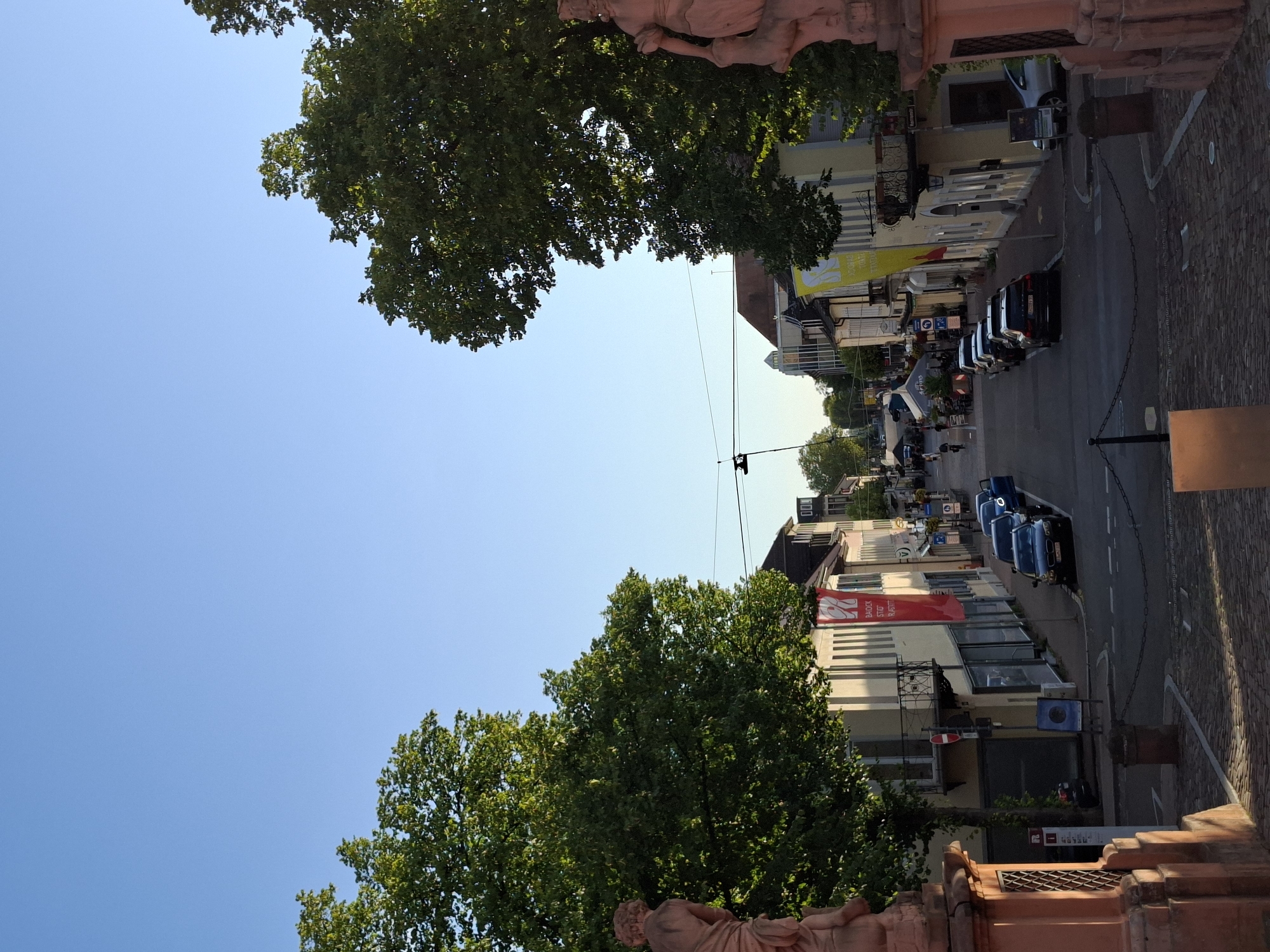 The image shows a street scene in Rastatt, Germany, likely taken from an elevated position, possibly a bridge. The street slopes downwards and is lined with buildings on both sides, featuring shops and balconies. Several cars are parked along the street. Large trees with green foliage flank the sides of the street, and a clear blue sky dominates the upper portion of the image. In the foreground, there are two large stone sculptures on either side, and a paved walkway with decorative chains. The overall impression is that of a well-maintained street in a European city, bathed in natural light, suggesting it is daytime, possibly late afternoon due to the warm lighting. There are a few pedestrians visible in the distance, walking along the street. The scene appears peaceful and ordinary.