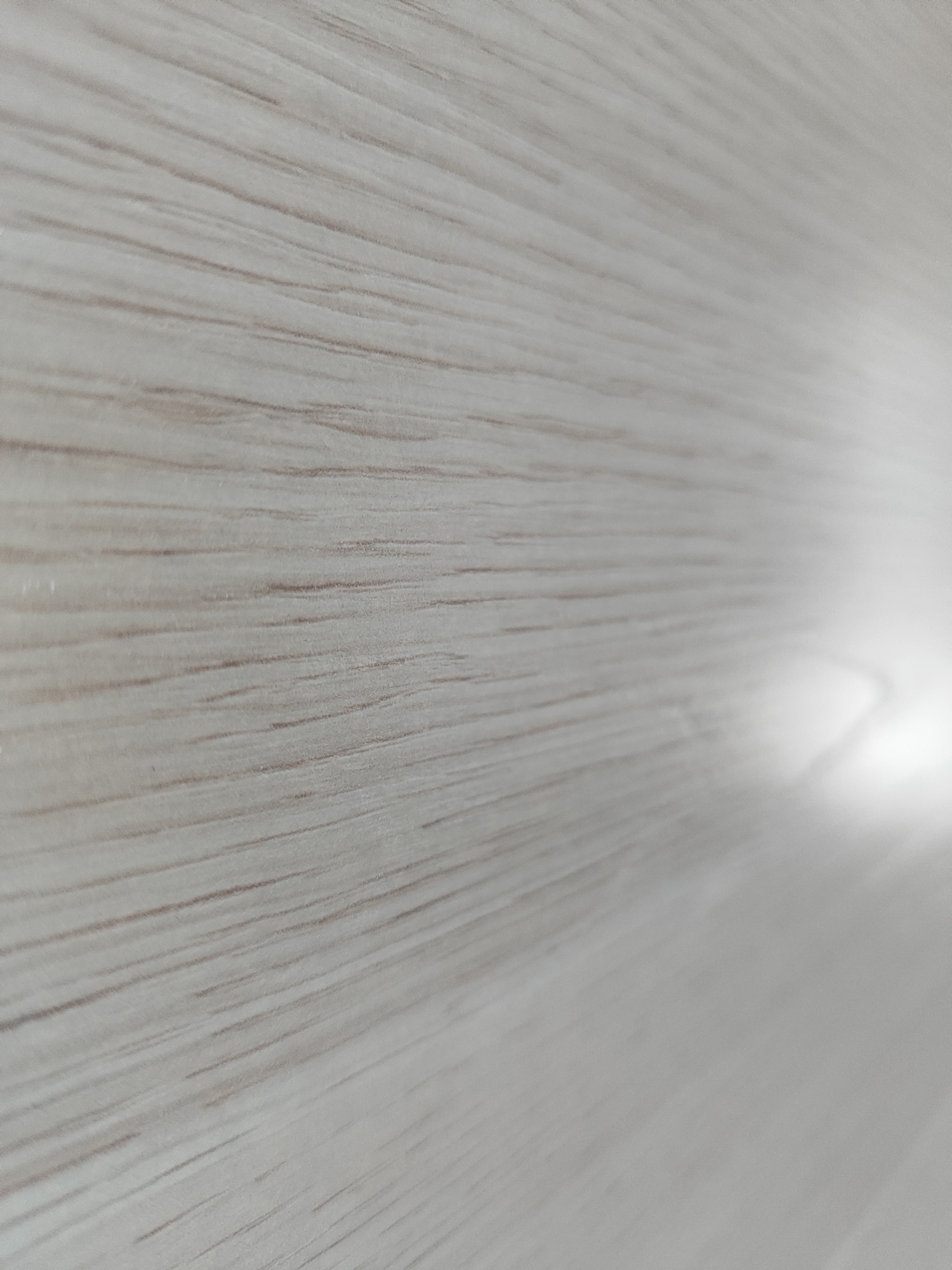 The image is a close-up, angled shot of a light-colored, textured surface, likely wood grain or a laminate material. The texture consists of faint, parallel lines running diagonally from the upper left to the lower right of the frame. The lighting creates a bright, blown-out area on the right side of the image, suggesting a light source is positioned there and out of frame. There are no people, specific objects, or actions depicted. The image appears to be a detail shot of a surface, not a scene within a city. Therefore, its location in Marusino, Russia cannot be determined from the visual information provided.