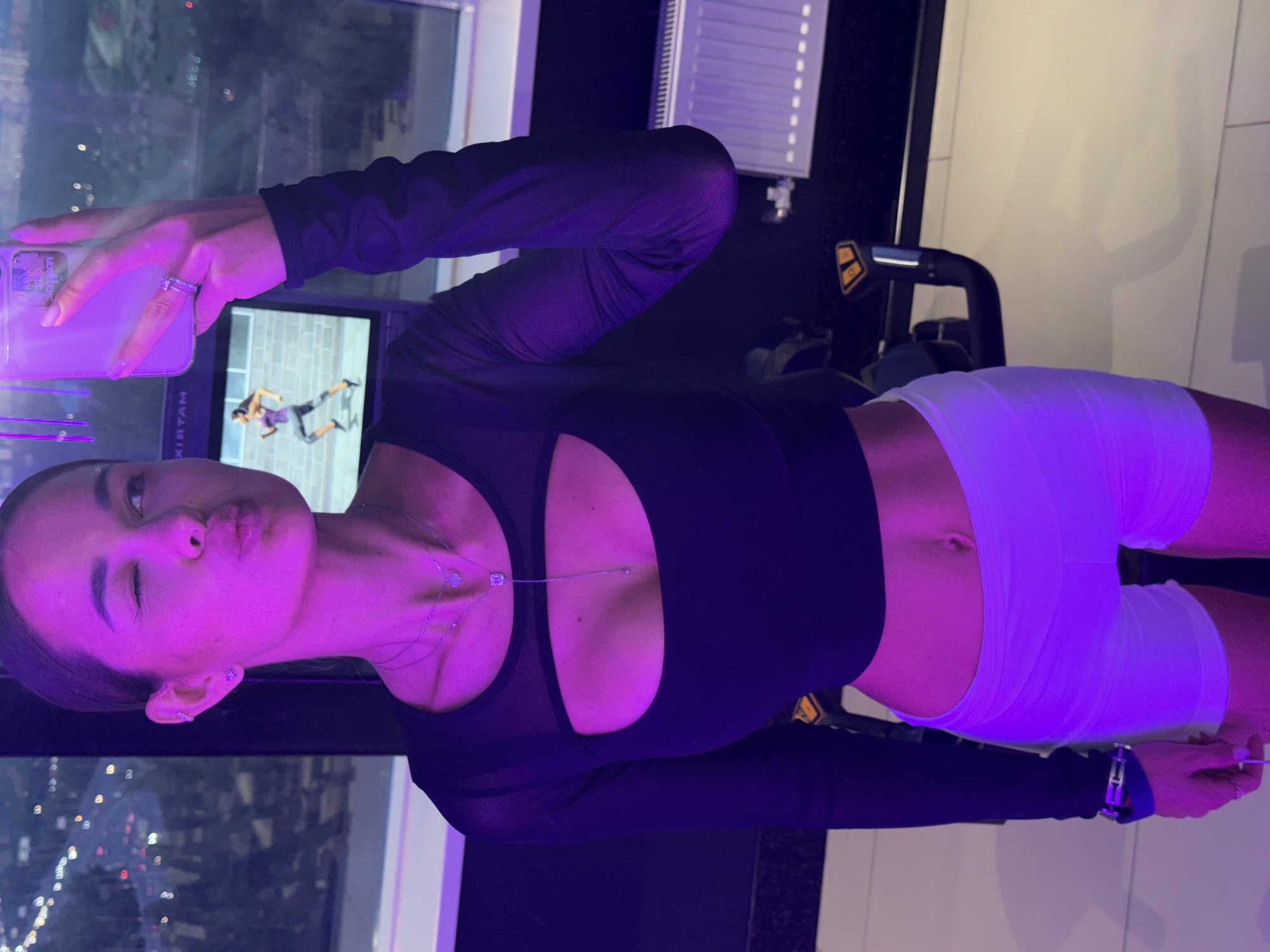 A young woman is taking a selfie in front of a window overlooking a city skyline. She is wearing a black long-sleeved top with a cutout in the chest area and white shorts. She is holding a smartphone in her left hand to take the picture. The city skyline is visible through the window. There is gym equipment in the background.
The location is not identified as Krasnoyarsk, Russia.