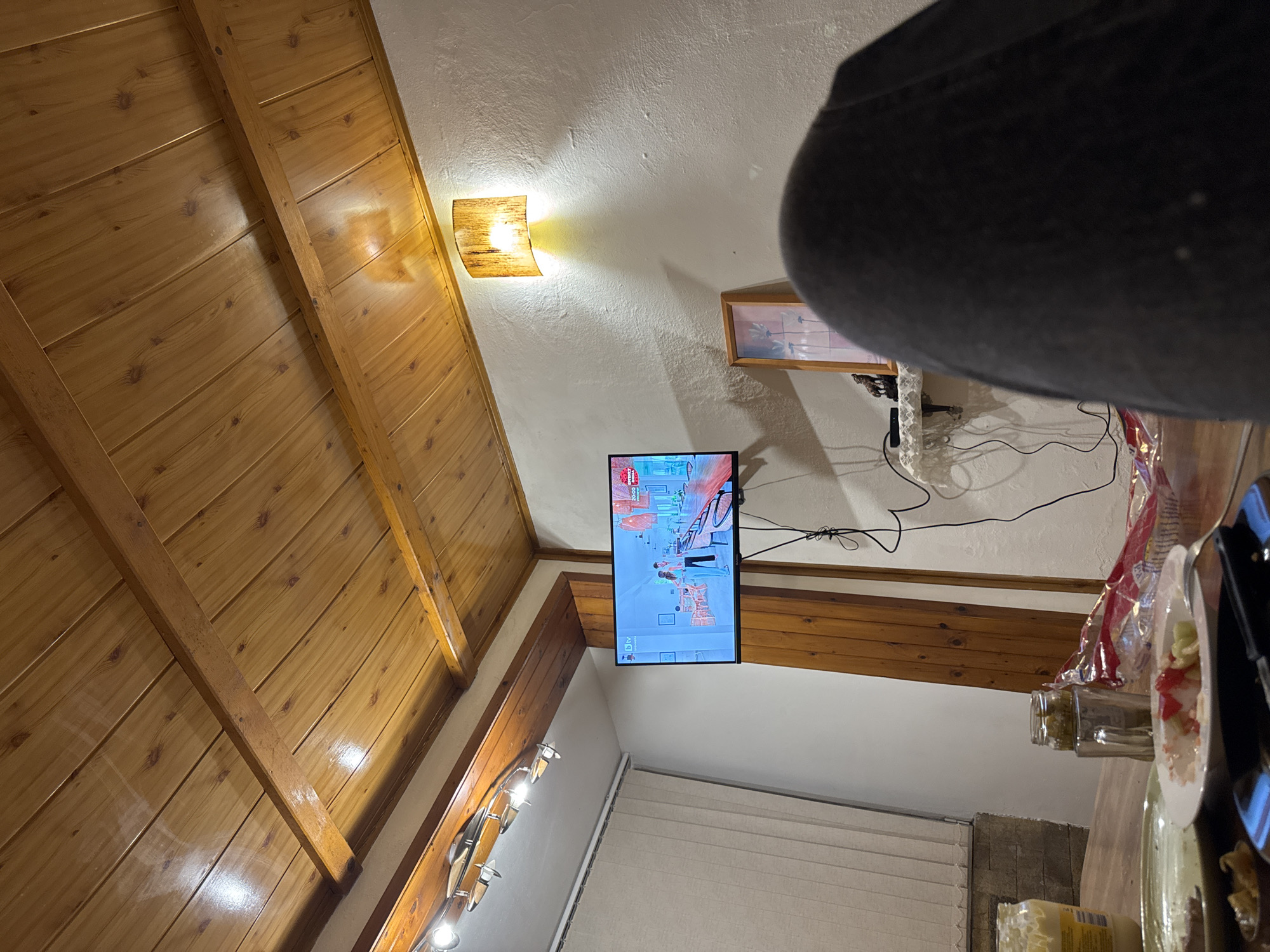 The image shows an interior space with a slanted, wood-paneled wall/ceiling on the left. A television is mounted on the wall, displaying a show. A wall sconce is located near the top of the wall. On the right side of the image is a partial view of a table with food items, including plates and jars. In the upper-right corner, a portion of a person's head is visible, likely the photographer. The background includes a white ceiling and a wall with light fixtures. The location is not apparent from the image.