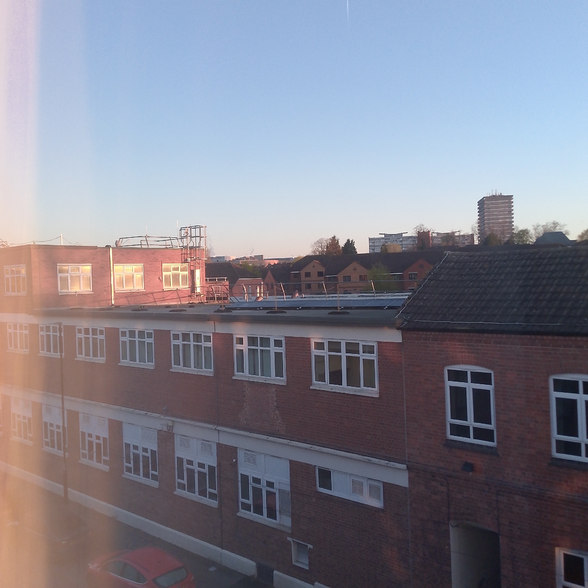 The photograph depicts a three-story brick building with white-framed windows, possibly an educational or institutional building, viewed from an elevated position. On the roof are some solar panels.
In the background, a cluster of residential houses with varied rooflines is visible, along with a tall high-rise building, potentially a residential tower. The sky is clear, with a gradient from light blue to a faint orange near the horizon, suggesting either sunrise or sunset. The scene gives the impression of the late afternoon or early morning in a urban area.
The image includes a red car parked in front of the building.
Based on visual landmarks, the buildings are similar to buildings in Coventry, United Kingdom.