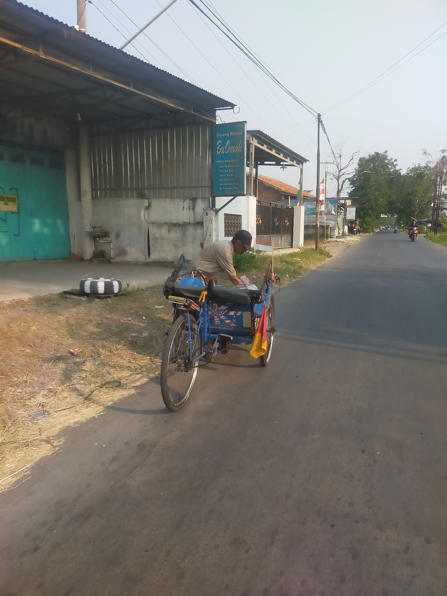 A man is seen from behind, wearing a beige shirt and a dark baseball cap, standing next to a blue and yellow pedicab on the side of a paved road in Kambangan, Indonesia. He appears to be arranging items in the pedicab. The pedicab has a black seat and a blue body with some white lettering. A small Indonesian flag is attached to the front of the pedicab. In the background, there is a building with a corrugated metal roof, a turquoise garage door, and a sign that reads "Warung Makan Bu Oman". Further down the road, several motorcycles are visible. The environment suggests a rural or semi-urban setting with dry grass and some trees. The sky is clear and light blue, indicating daytime.