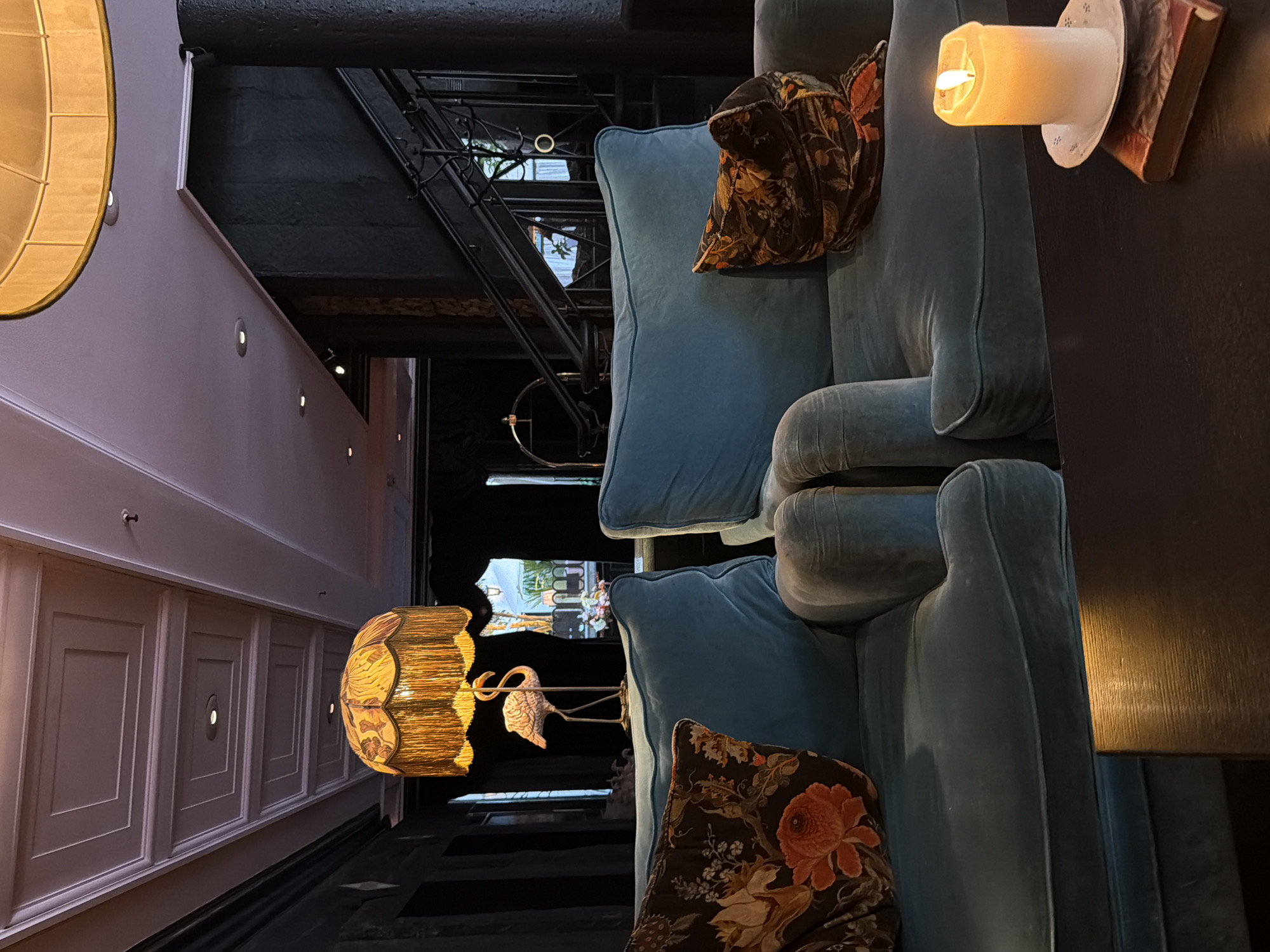 This image depicts the interior of a dimly lit, upscale lounge area in Malmö, Sweden. The foreground is dominated by two plush, teal-colored armchairs, each adorned with a floral-patterned cushion. To the right, a lit white candle rests on a small decorative plate atop a closed book, casting a warm glow. In the background, a ornate lampshade with fringed edges hangs over a decorative flamingo statue. A dark staircase with a wrought-iron railing ascends on the right side of the frame. The ceiling features a coffered design with recessed lighting. The overall ambiance is one of sophisticated relaxation.