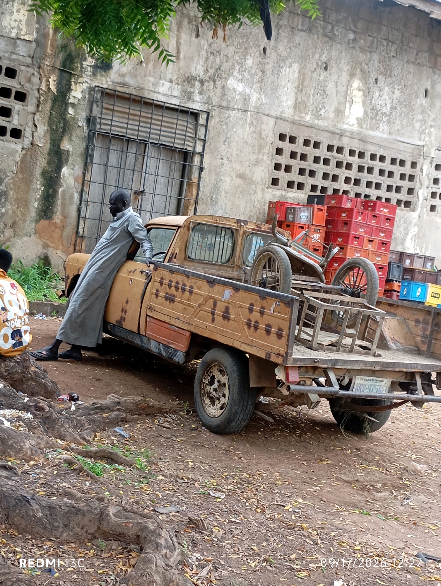 A man in a gray robe leans against the driver's side door of a weathered, rusty yellow pickup truck. The truck bed is filled with stacked red and black crates, and a dismantled cart with two wheels. In the background, a dilapidated concrete building with a barred window and square ventilation holes stands. The ground is dusty and covered with fallen leaves and debris. Another person, partially visible in the lower left, wears a yellow shirt with cartoon characters. The license plate of the truck reads "NASARAWA KRU-9754EZ". The scene appears to be in Shendam, Nigeria.