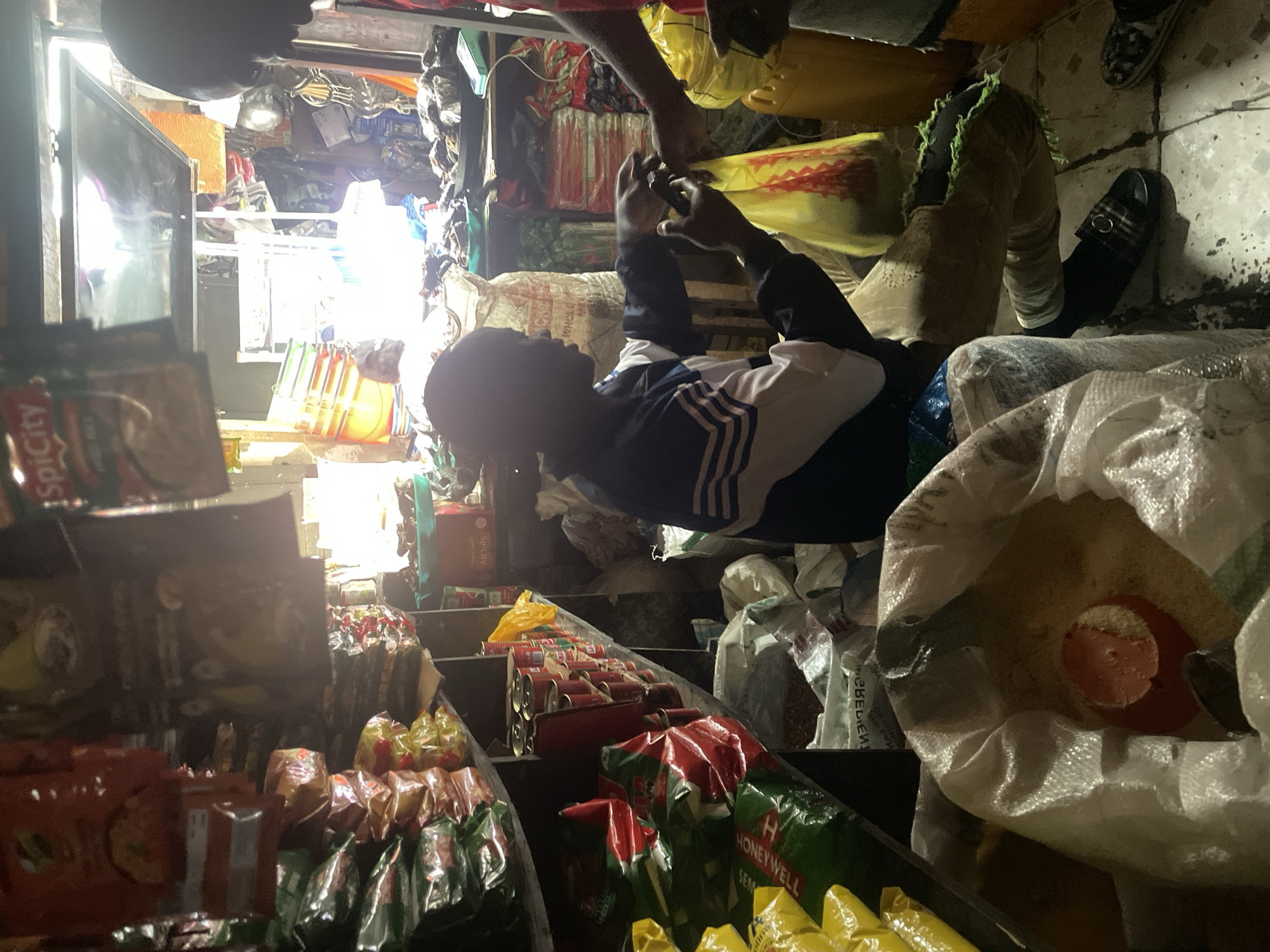 The image depicts a scene inside a small shop, likely a market stall, in Abuja, Nigeria. The interior is densely stocked with various goods, suggesting a retail environment. In the foreground, a person is seated, holding a yellow plastic bag, and appears to be interacting with another individual whose hand is reaching into the bag. The seated individual is wearing a dark jacket with white and blue stripes on the sleeves and chest. Numerous sacks and packaged goods are visible on shelves and the floor, including what appears to be rice in a large white sack in the lower left of the frame. The shelves are laden with canned goods, plastic-wrapped food items, and other bagged products. The lighting is dim and appears to be coming from the back of the shop, creating shadows and highlights. The overall atmosphere suggests a busy, functional retail space.