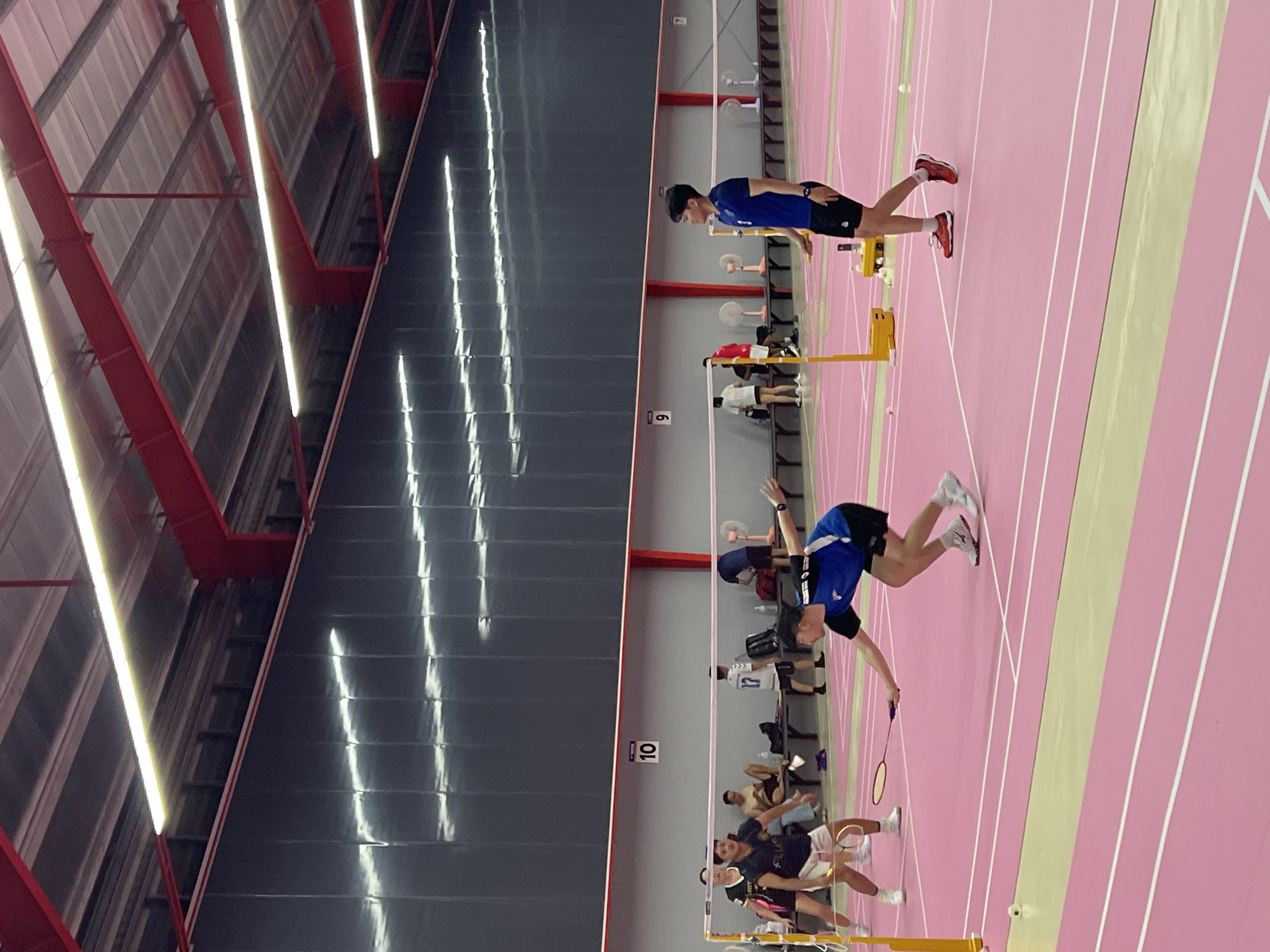 The image shows a badminton match taking place indoors on a pink court with white lane markings. Several people are visible, some actively playing and others spectating. In the foreground, two male athletes are engaged in a rally. One is in mid-air, reaching for a shot with a badminton racket, wearing a blue and black outfit. The other is in a lower stance, preparing to receive the shot, dressed in a black and white outfit. The background shows spectators seated and standing along the side of the court. The lighting appears to be artificial, suggesting an indoor facility. The ceiling has a metallic structure with red beams and long fluorescent lights. There is a sign with the number "10" visible on the wall. The setting is a sports hall, likely a gymnasium, designed for athletic competitions. There is no indication of the time of day or weather conditions as it is an indoor scene. The overall atmosphere seems to be one of active sport.