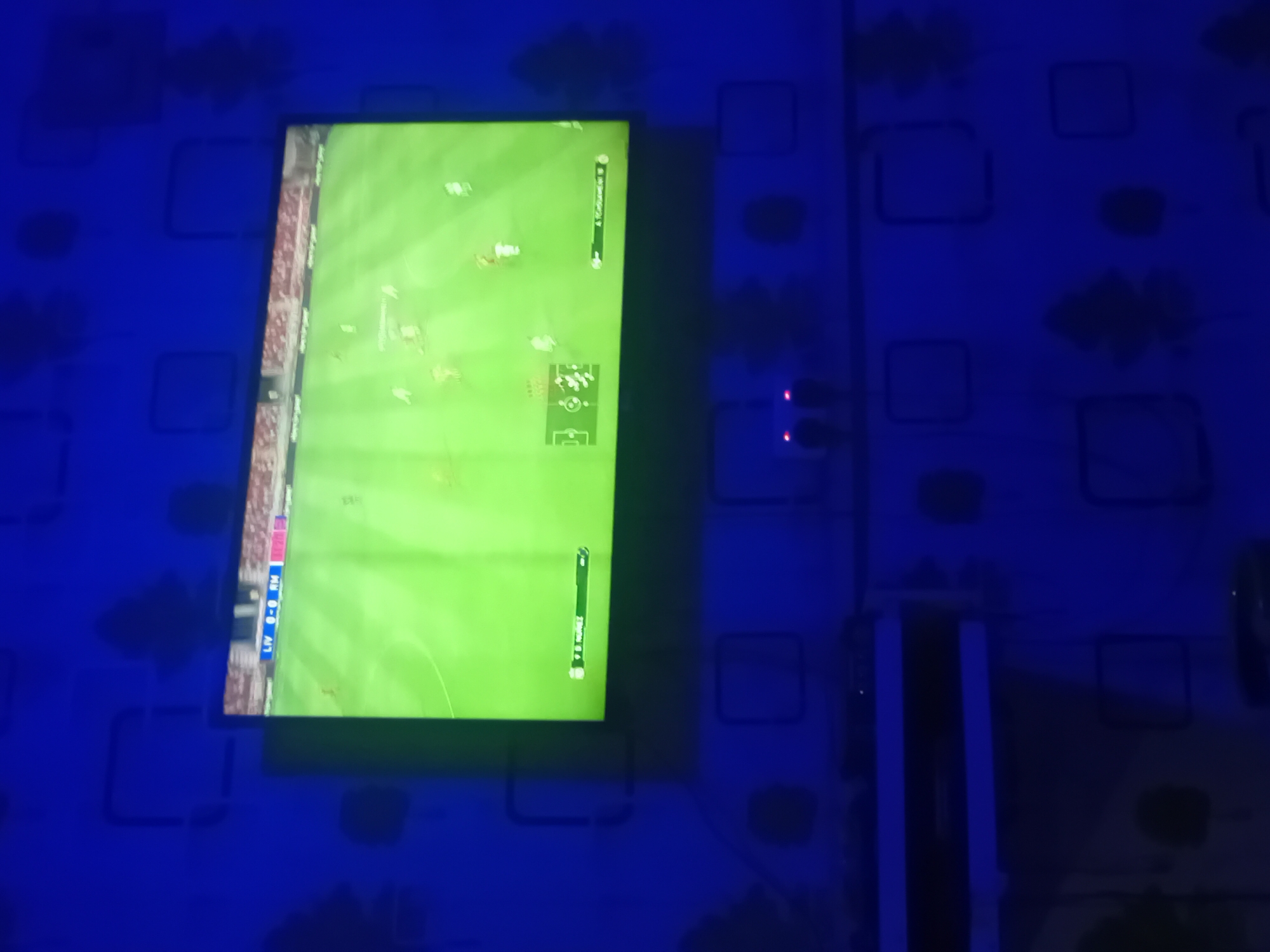 This media file captures an indoor scene, likely a gaming setup, under strong blue lighting. The primary subject is a television screen mounted vertically on a dark blue wall. The wall features a repeating pattern of lighter blue square and amorphous shapes, possibly from wallpaper or a painted design.
The television screen displays a football (soccer) video game, with players visible on a green pitch. In the bottom left corner of the screen, the scoreline "LIV 0 - 0 MN" is visible, suggesting a match between Liverpool and another team, possibly Manchester United. On the top right, what appears to be a player's username, "@Criggeland," is displayed. The bottom right of the screen shows "2:08," likely an in-game timer, and "Trons 11," possibly a team or player name.
To the right of the television, against the blue wall, several electronic devices are visible. Two small black objects with red indicator lights are present, likely power adapters or similar peripherals. Further down and to the right, a larger rectangular object, possibly a gaming console, is positioned, also featuring red indicator lights.
The ambient lighting is distinctly blue, casting a strong hue across the entire scene and creating a dark, atmospheric setting, indicative of a room with controlled lighting, likely at night. The vertical orientation of the television suggests a non-traditional setup, possibly for a specific gaming experience or to fit the space. The location context provided is Lokoja, Nigeria, though no specific visual cues within the image confirm this.