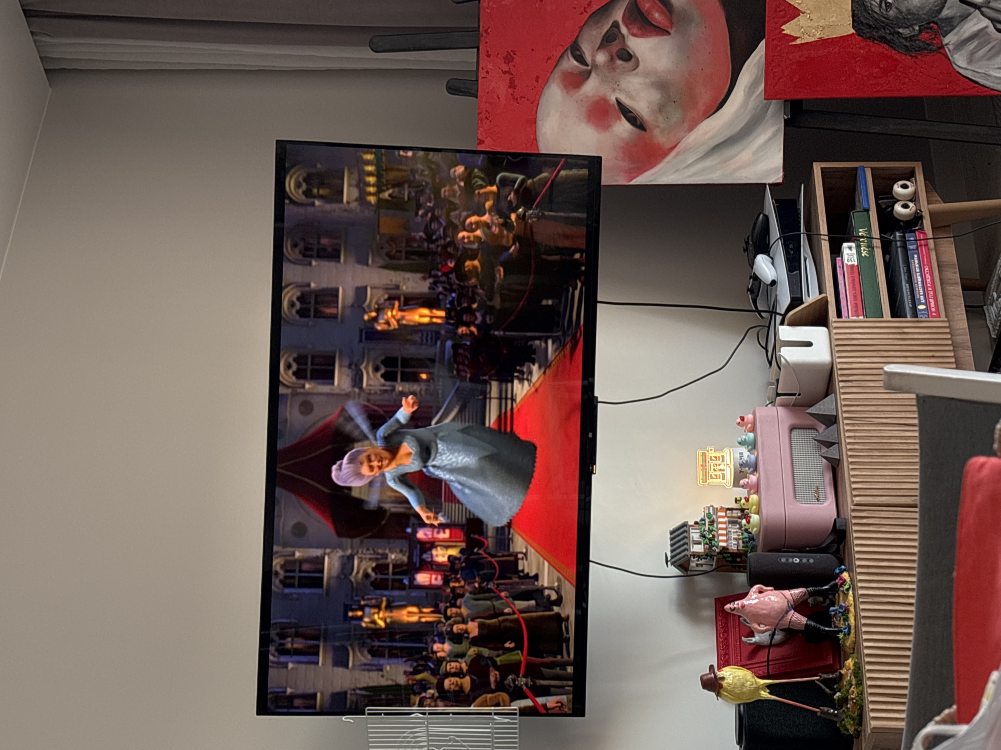 The image captures an indoor scene, likely a living space in Krasnoyarsk, Russia, featuring a television, artworks, and a decorative shelving unit.
The central focus is a large, black flat-screen television displaying an animated scene. A character resembling the Fairy Godmother, with purple hair and a blue dress, is shown mid-action on a red carpet, flanked by a crowd of animated figures and ornate buildings. This appears to be a scene from an animated movie, possibly *Shrek 2*.
Above and to the right of the television, two framed artworks are visible. The left painting features a close-up of a person's face with pale skin, flushed cheeks, and red lips against a vibrant red background, partially covered by white drapery. The right painting, partially obscured, also uses a red background and shows a portion of a face with dark lines and a golden, crown-like element.
To the right of the television, a light wooden, multi-compartment shelf holds various items. The top section contains several books, including one green-spined book labeled "VETRUSE" and another with a red spine featuring Russian text, "Учебное пособие по истории древнего мира" (Textbook on the history of the ancient world). Two decorative white eyeballs on sticks are also placed here, along with a pen.
Further down the shelf are several decorative objects and electronic devices. These include a pink portable speaker, a black cylindrical speaker, a small house model with a glowing sign, multiple small pig figurines, a yellow bird figurine wearing a hat, and a whimsical pink pig-like figure dressed in fishnet stockings. A white container with a cutout for cables sits on the shelf below the TV.
The wall is a light, neutral color. Black cables are visible, connecting various electronic devices. The overall impression is a cozy, personalized interior space filled with entertainment and decorative elements.