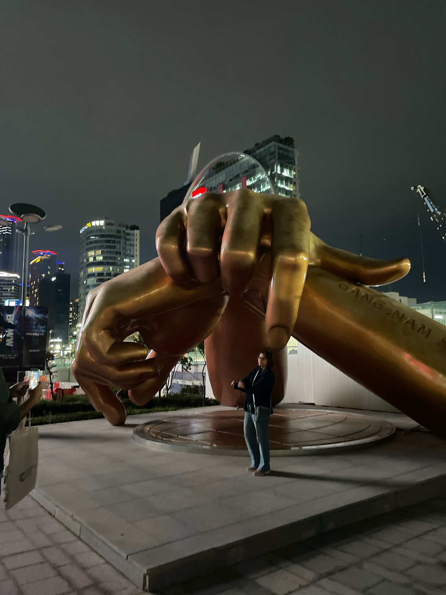 This image depicts the "Gangnam Style" statue located in front of COEX East Gate in Seoul, South Korea. The statue, a large golden sculpture of two hands forming a gesture from the famous music video, is the central focus of the scene. A woman wearing a dark jacket and light blue jeans stands in front of the statue, appearing to pose or observe. To the left, partially visible, a person in a green shirt holds a white tote bag and takes a picture with a phone. The background reveals illuminated skyscrapers and city lights, indicating an urban setting at night. A crane is also visible in the far right background. The ground is paved with concrete slabs.