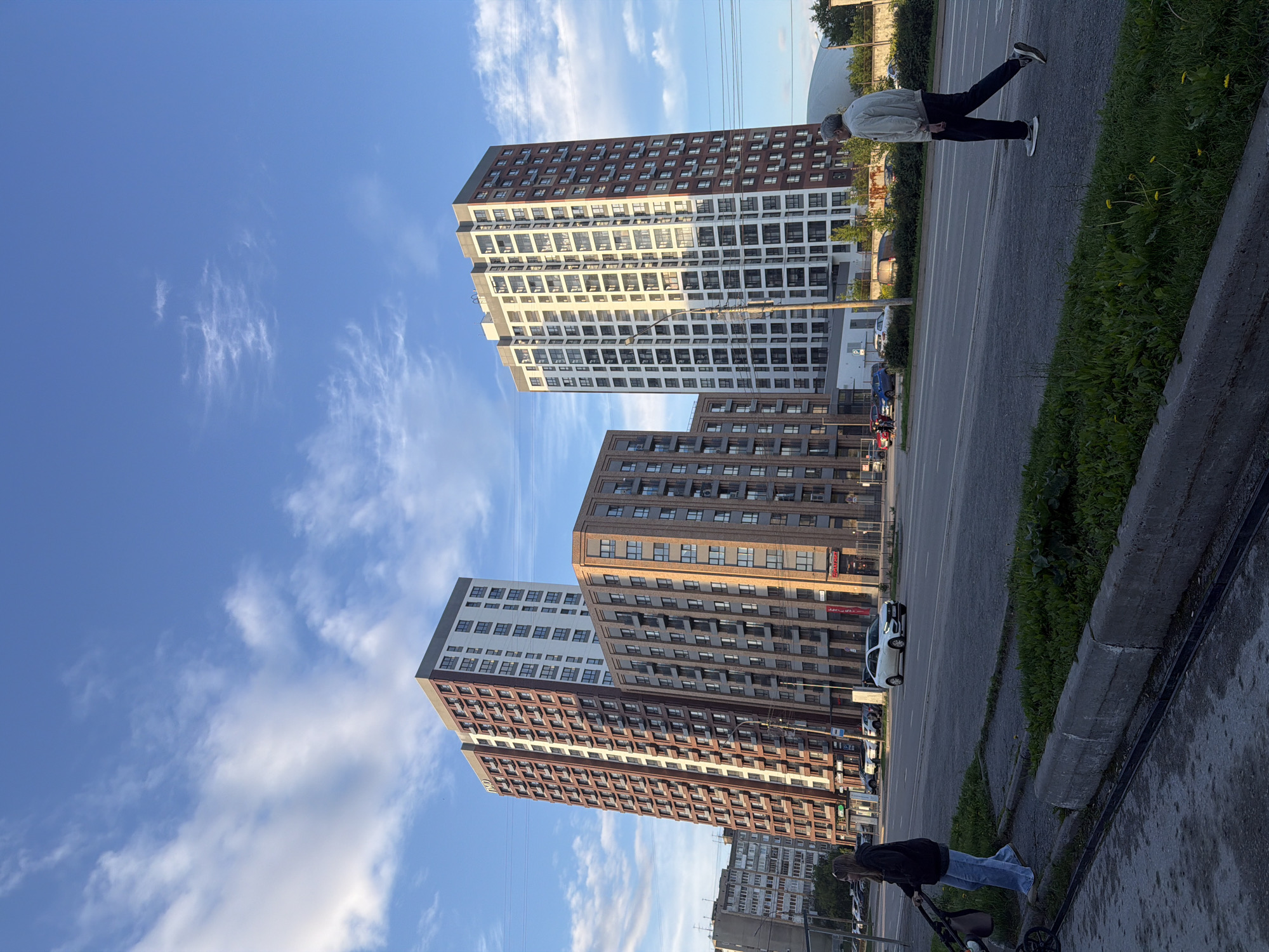 This is a low-angle, medium shot of a city street in Perm, Russia, on a clear day with a few scattered clouds. The focus is on modern apartment buildings. In the foreground, a paved sidewalk and road are visible, with a concrete curb and some grass. Two people are present: a woman is pushing a stroller on the left side of the sidewalk, and an older man in a light-colored jacket and dark pants is walking on the right side of the road, facing away from the camera.
Three tall, modern apartment buildings dominate the background. The building on the far left is a tall, slender tower with a reddish-brown facade and white accents, featuring many balconies. To its right is a shorter, wider building with a reddish-brown brick facade and numerous windows. The tallest building, on the far right, is a white tower with dark accents and many windows, also with numerous balconies. In the distance, other buildings and some trees are visible, along with power lines stretching across the sky. The overall impression is one of contemporary urban development.