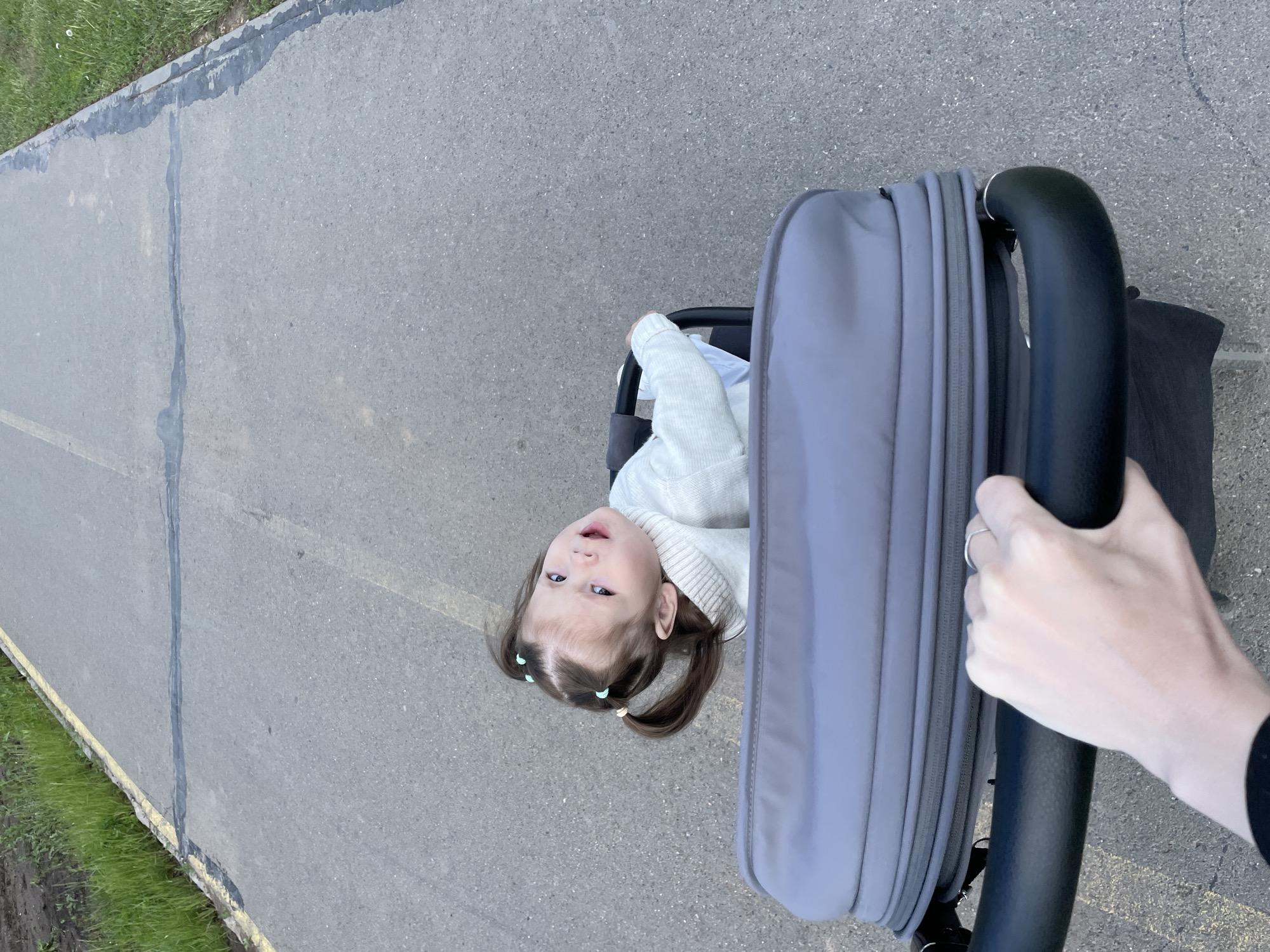 A child is in a stroller on an asphalt path next to a grassy area in Izhevsk, Russia. The path has yellow lines on one side and patches of sealant in cracks. The child, wearing a light-colored sweater, has pigtails secured with hair ties. A hand holds the stroller's handle.