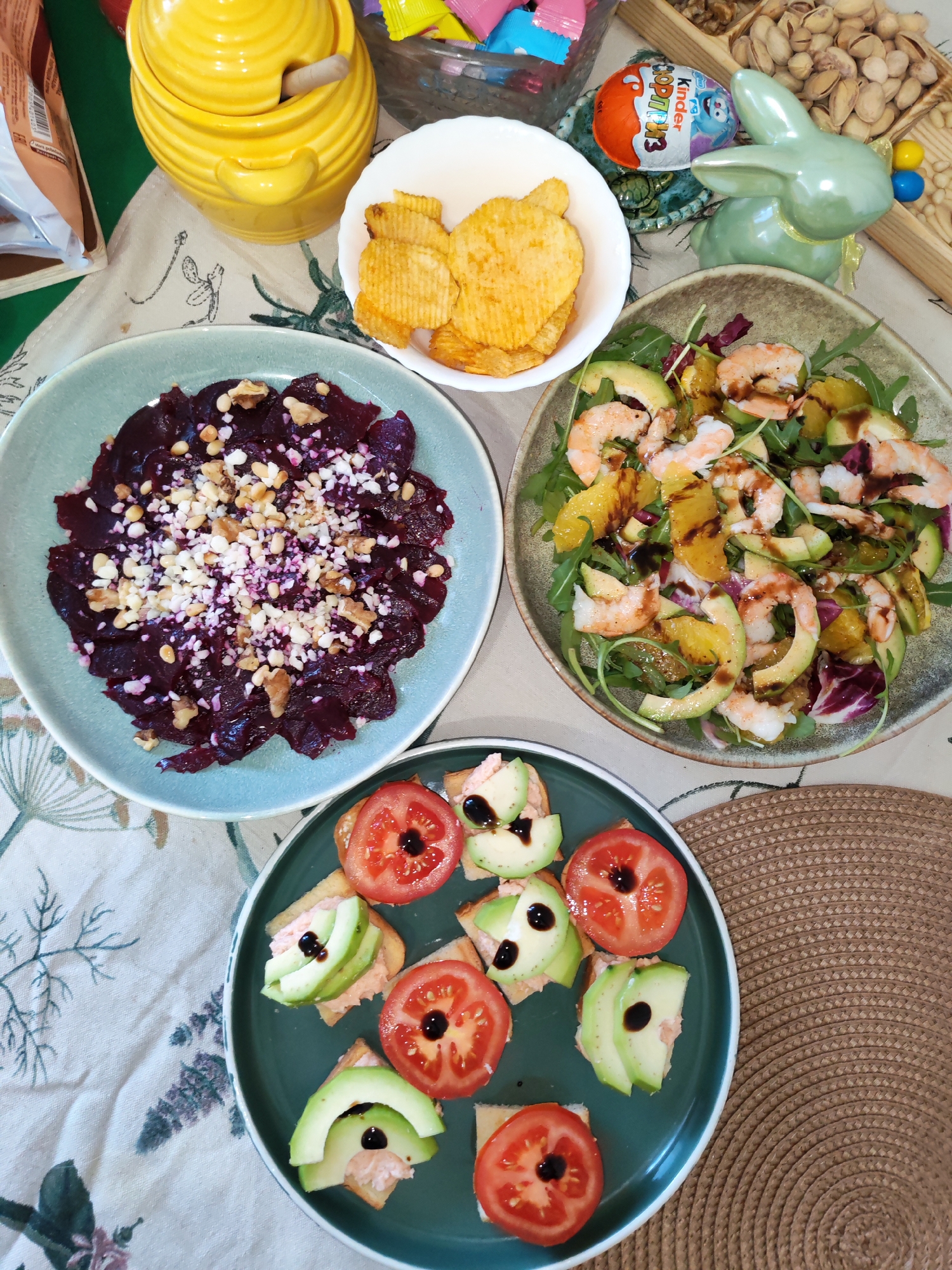 An overhead shot displays a vibrant table laden with various dishes and snacks, suggesting a casual gathering or meal. The table is covered with a white tablecloth featuring subtle green and purple floral or botanical patterns.
In the center-left, a light blue-green ceramic bowl holds thinly sliced beetroot, possibly a carpaccio, generously topped with chopped walnuts and a white, crumbly ingredient, likely cheese or finely ground nuts. To its right, a white bowl contains crinkle-cut potato chips. Further right, a larger bowl with an earth-toned rim presents a colorful salad composed of peeled shrimp, slices of avocado, orange segments, leafy greens (including arugula and possibly radicchio), all drizzled with a dark balsamic glaze.
At the bottom, a dark green ceramic plate features an arrangement of small appetizers or canapés on bread squares. Some are topped with a slice of red tomato and a dot of dark glaze, while others have a pink spread (possibly fish or seafood based) topped with avocado slices and a similar dark glaze. A circular woven placemat is visible under this plate on the right.
Additional items on the table include a bright yellow ceramic honey pot with a wooden dipper at the top left. A clear bowl of assorted colorful candies is nestled behind it. To the right of the chips, a wooden tray holds shelled pistachios and walnuts. A light green ceramic bunny figurine with a yellow ribbon is positioned next to the nuts, and a Kinder Surprise egg featuring the text "KINDER СЮРПРИЗ" (SURPRISE) is visible nearby, adding a festive, possibly Easter-themed, touch. A partially visible snack bag is in the background on the far left. The scene evokes a relaxed, celebratory atmosphere in Izhevsk, Russia.