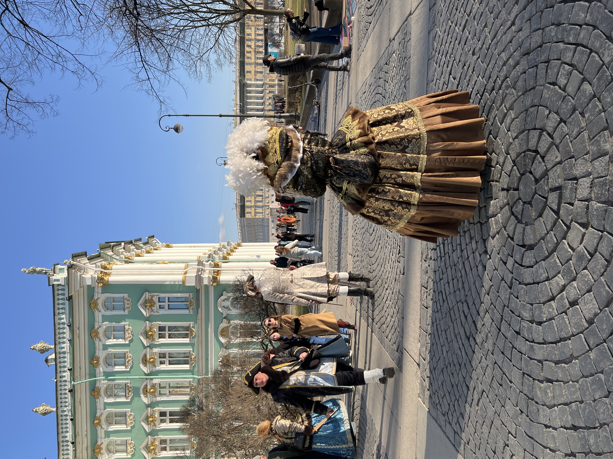 The image depicts an outdoor daytime scene on Palace Square in Saint Petersburg, Russia, under a clear blue sky. The prominent building in the left background is the light green and white Winter Palace, part of the Hermitage Museum, characterized by its ornate Baroque architecture with gold accents, white columns, and roofline statuary.
The ground is paved with dark grey cobblestones arranged in a distinctive concentric and radial pattern. In the immediate foreground, a person is seen from the back, wearing an elaborate historical costume featuring a voluminous brown and gold patterned brocade gown, a pleated brown underskirt, a fur-trimmed bodice, and a large white feathered headdress. A small patterned drawstring bag hangs from the back of the costume.
To the left in the mid-ground, a person walks towards the left, dressed in a black and gold historical coat over a light blue vest, dark trousers, white leg wraps, and a black tricorn hat. Partially obscured behind them is another individual in a blue and gold historical dress with a fur-trimmed hat. To the right of the central costumed figure, another person in a light-colored, patterned historical coat walks away from the viewer, wearing dark pants and black boots with white cuffs.
A black, ornate street lamp stands in the mid-ground. Numerous other individuals, some in modern clothing and others in historical attire, populate the square in the background. On the right side, a person in a black puffer jacket and dark pants stands upright, observing the scene. Further right, another person in a dark jacket and blue pants kneels on the pavement, interacting with colorful items laid out on the ground. Bare tree branches are visible against the sky on the right. In the far background, across the square, a large yellow neoclassical building with white columns, consistent with the General Staff Building, is visible.