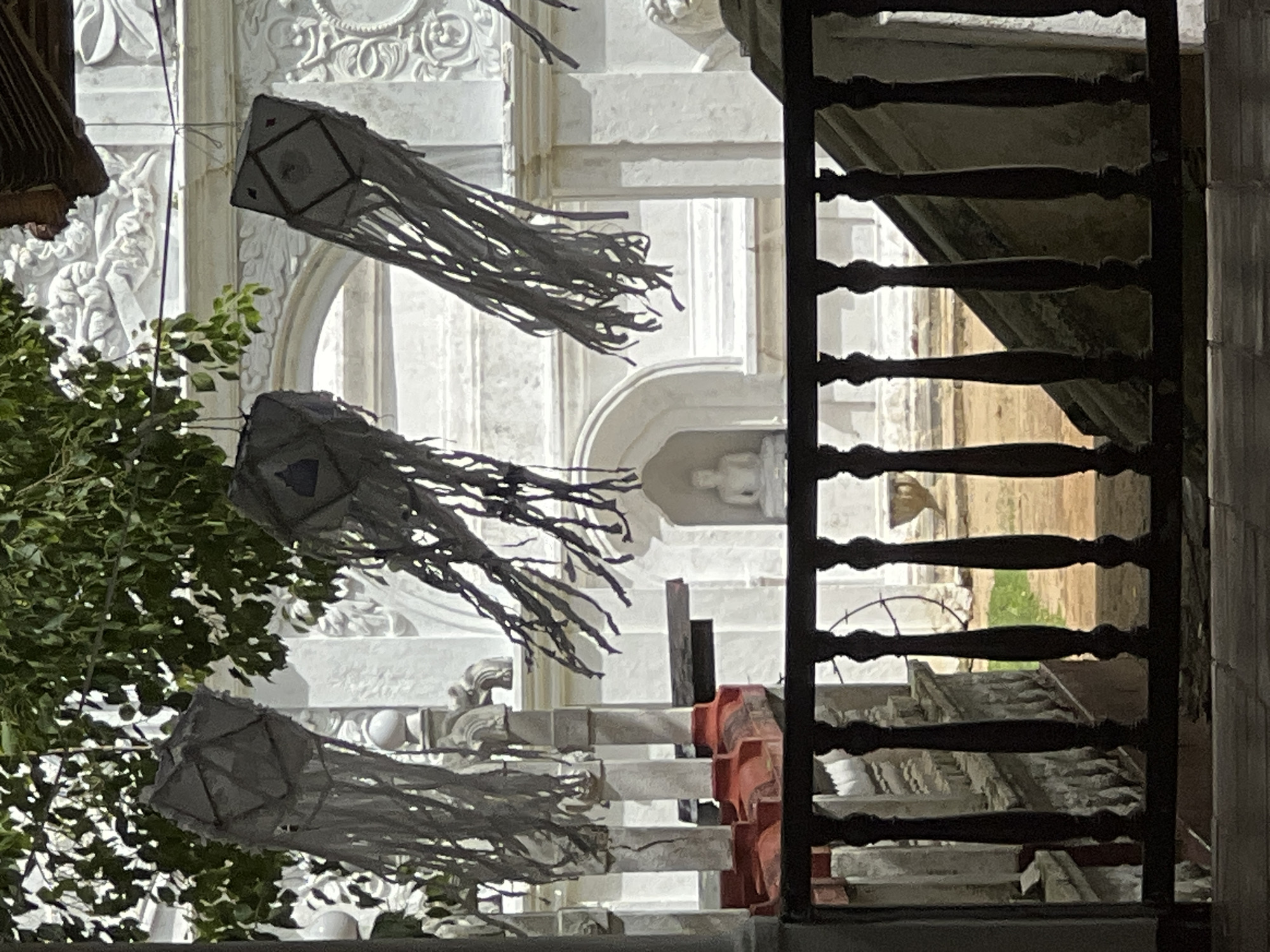 The image displays a vertical composition featuring decorative lanterns and green foliage set against a backdrop of ornate white architecture, framed by a dark railing.
In the foreground and mid-ground, three prominent decorative lanterns are suspended. Each lantern has a geometric top section, possibly hexagonal or square, from which long, fringed streamers cascade downwards. They appear to be made of a light-colored material, likely white or pale grey. To the left, vibrant green foliage from a tree or bush partially obscures the view. On the right side, a dark, ornate railing or balustrade with turned spindles creates a strong vertical element and provides a stark contrast to the bright background.
The background is dominated by a grand, white architectural structure, likely a temple or a building of cultural significance. This structure is adorned with intricate carvings, including floral motifs and possibly figures, along with arches and columns. Within one of the arched recesses, a small, light-colored statue, possibly a Buddha, is discernible, reinforcing the cultural context. Additional architectural elements such as ledges, steps, and balustrades are visible, with some showing signs of age or wear. Partially visible red or orange objects in the mid-ground may be temporary barriers or construction materials.
The lighting highlights the bright white surfaces of the building, creating deep shadows on the darker elements like the railing and foliage. This suggests an outdoor setting during the daytime with strong light, or possibly early evening with artificial illumination. The presence of the lanterns strongly implies a festive occasion, a cultural celebration, or a religious event. No people or visible text are present in the image. The location cues, particularly the architectural style and lanterns, suggest a South or Southeast Asian setting.