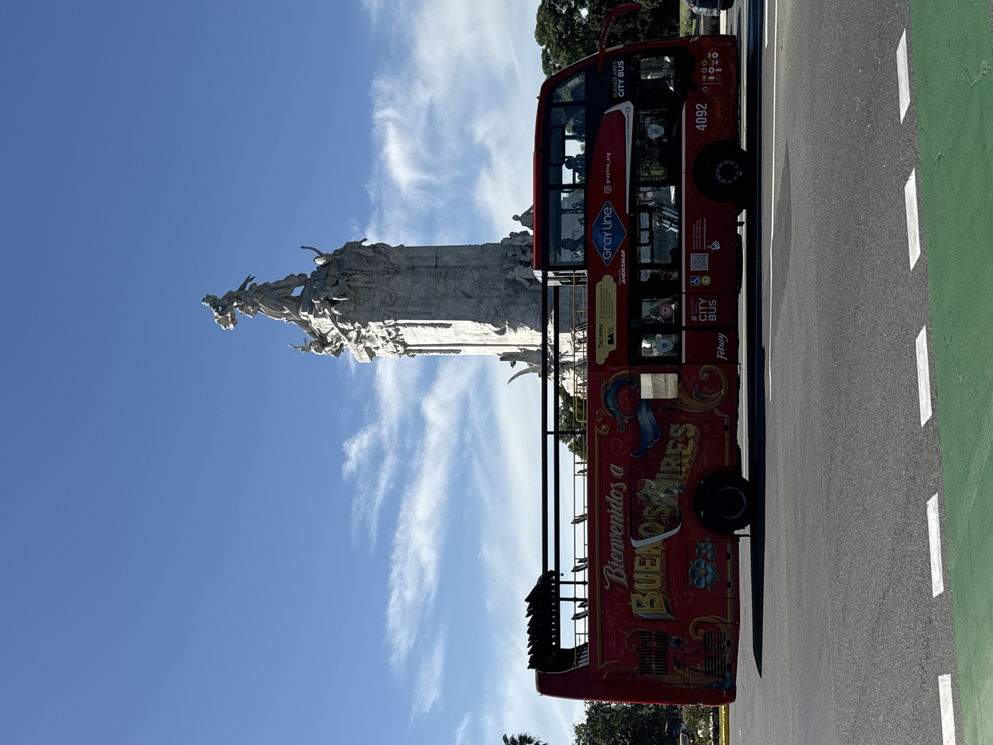 The image, correctly oriented, captures a sunny daytime scene in Buenos Aires, Argentina, featuring a large stone monument and a red double-decker city tour bus.
The prominent subject on the left is an ornate, multi-tiered white stone monument, possibly a memorial or obelisk, richly adorned with numerous sculpted figures. These figures appear to be in various poses, some reaching upwards, contributing to the monument's grand and historic aesthetic. The upper part of the monument is silhouetted against a vibrant, clear blue sky with some wispy white clouds.
On the right, a red "BUENOS AIRES CITY BUS" double-decker tour bus is seen driving on a paved road. The bus features a classic design with an open-top upper deck, where several passengers are visible, suggesting a sightseeing tour. The side of the bus is brightly decorated with "Bienvenidos a BUENOS AIRES" (Welcome to Buenos Aires) in stylized lettering, along with the "Gray Line" logo, "ANDESMAR," "Turismo BA," and the bus number "4092." Other smaller texts like "#BUENOS," "CITY BUS," and "Febuey" are also visible, along with an Instagram handle "grayline_arg."
The bus is traveling on a road marked with white dashed lines and a distinct green lane on the far right, possibly for bicycles or a dedicated bus lane. Lush green trees are visible in the background, peeking from behind the bus and monument, suggesting a park or treelined street. The bright sunlight and clear sky indicate pleasant weather, ideal for outdoor touring. The overall impression is one of active tourism in a beautiful city setting.