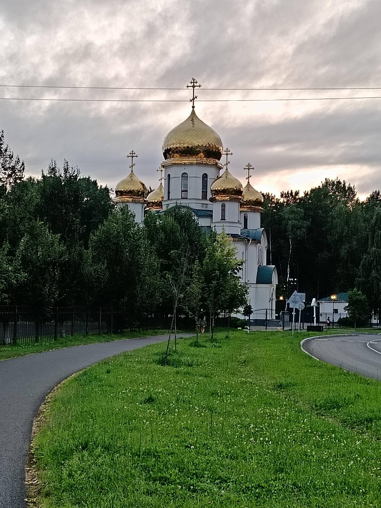 The image shows a white Orthodox church with multiple golden domes, set against a backdrop of lush green trees and a cloudy sky. In the foreground, a paved pathway curves around a grassy area dotted with small white flowers. A black metal fence runs along the left side of the pathway. A road is visible on the right side of the image. The overall scene suggests a serene and possibly late afternoon setting in a park-like area within a city.