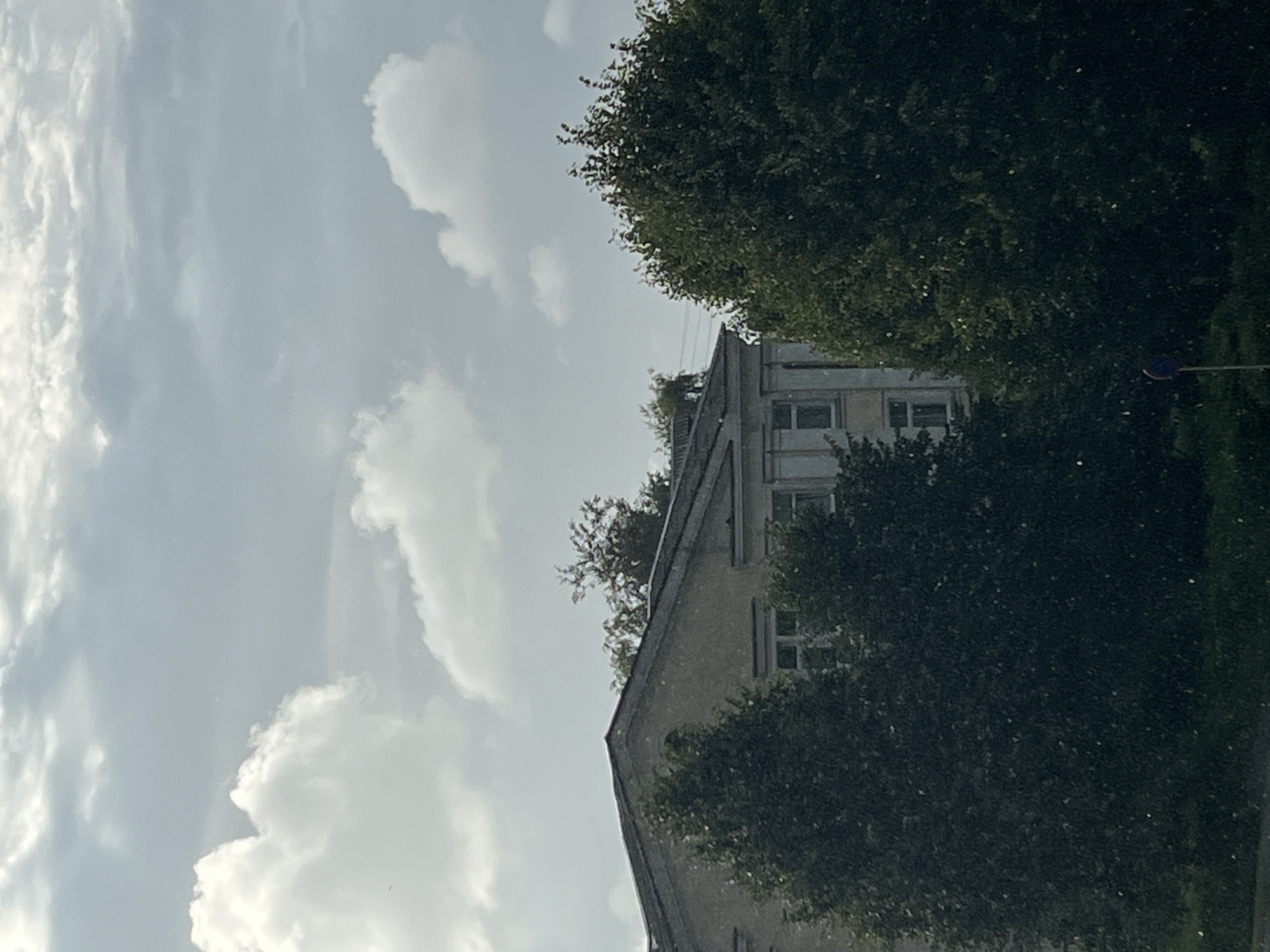 The image depicts a building with a gray roof and light-colored facade, partially obscured by lush green trees. The sky above is overcast with a mix of white and gray clouds. The scene appears to be an outdoor urban environment in Kohtla-Järve linn, Estonia, possibly a residential or institutional building given its architecture. Small white specks are visible throughout the image, which could be rain, snow, or light glinting off airborne particles. In the foreground, dark green foliage dominates, with a hint of a traffic sign pole with a blue and red circular sign visible on the lower right side. The overall impression is a slightly hazy, cloudy day.