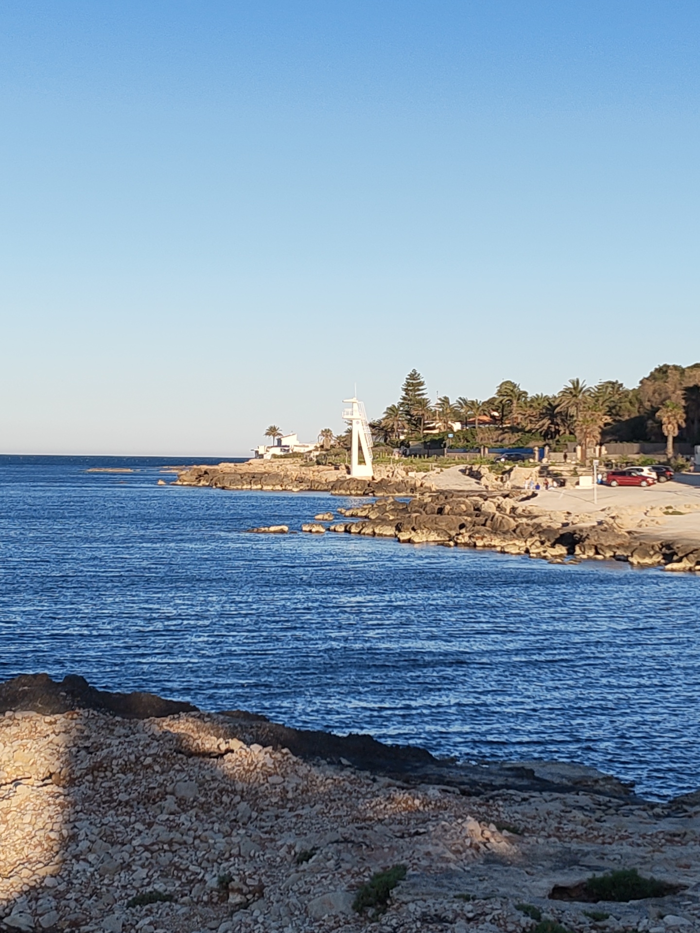 The image captures a serene coastal scene in Rotes, Spain, under a clear, bright blue sky. The deep blue sea, with gentle ripples visible on its surface, dominates the left and central parts of the frame. A rugged, dark rocky shoreline extends from the mid-ground, curving towards the right side of the image and into the distance.
On the prominent rocky outcrop, a distinctive white, A-frame or triangular structure, likely a diving platform or lifeguard tower, stands out against the green foliage behind it. Beyond the immediate shore, the land rises slightly, densely covered with various trees, including numerous palm trees, and some coniferous trees. Several light-colored buildings are nestled among the greenery.
Further to the right, a paved area or road runs parallel to the coast, where at least two cars, one red and one light-colored, are parked. A small group of indistinct figures, appearing to be 3-4 people, can be observed near the paved area.
The immediate foreground is characterized by rough, light-colored rocky terrain, partially bathed in sunlight and shadow. The lighting suggests either early morning or late afternoon, with clear, direct sunlight illuminating parts of the scene and casting soft shadows. The weather appears calm and sunny, indicating an inviting day for coastal activities. No visible text is present in the image.