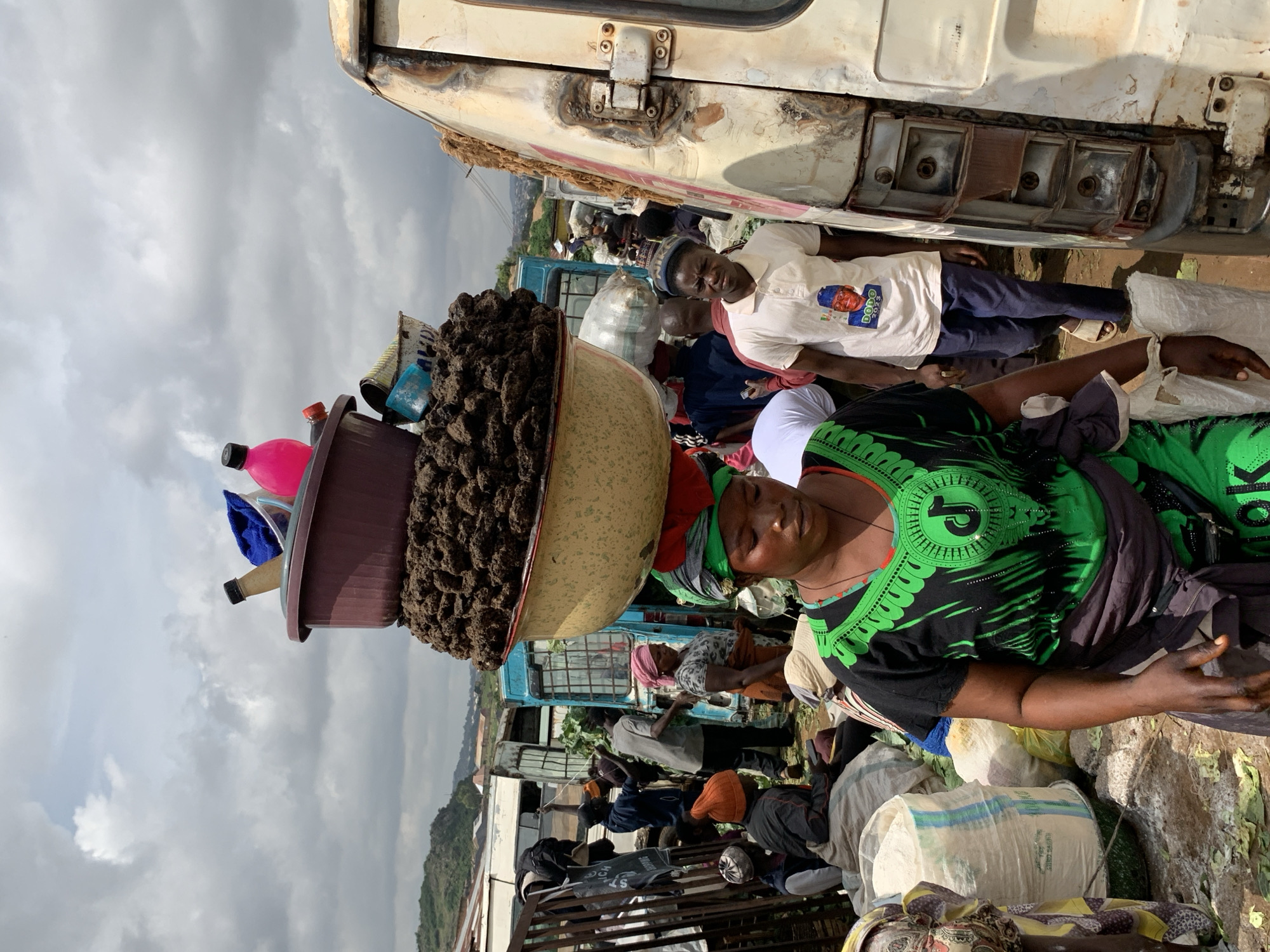 In Gura Top, Nigeria, a woman walks through a bustling market carrying a precarious load of items balanced on her head. The assortment includes multiple bowls, one large and brown, containing what appears to be dark, granular material, possibly soil or some form of produce. Beneath this, another bowl, this one yellow and rimmed with red, holds more of the dark material. Resting on top of the brown bowl are several plastic bottles, one bright pink, another clear with a black lid, and a blue cloth. The woman is dressed in a vibrant green and black garment with an intricate design, and a purple sash is wrapped around her waist. She is surrounded by other market-goers, some carrying goods and others appearing to be vendors or shoppers. In the background, several vehicles, including a white van with damage to its rear lights and a blue truck, suggest this is a busy transportation hub within the market. Bags of produce and other goods are visible, piled on the ground. The sky above is overcast with dramatic clouds. The scene captures a typical day of commerce and activity in this Nigerian city.