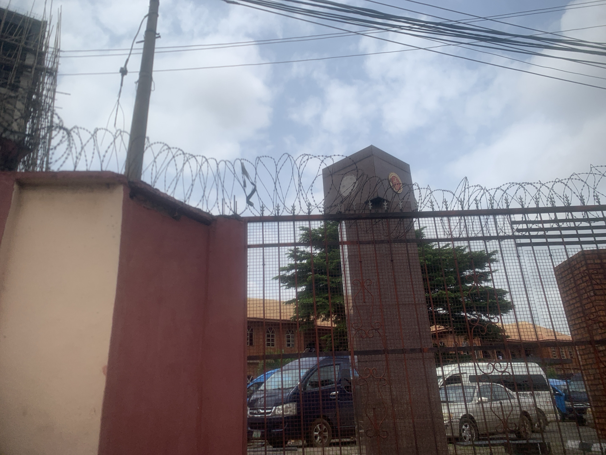 This image depicts a secured building entrance in Onitsha, Nigeria. A red and cream-colored wall with barbed wire atop it borders the foreground, leading to a metal gate. Beyond the gate, a dark blue van and a silver van are parked. In the background, a light brown building with a terracotta roof and arched windows is visible, along with several trees. A square, dark-colored pillar with insignia is also present near the gate, suggesting it is a landmark or part of the building's signage. Overhead, a tangle of electrical wires spans the sky, which is partly cloudy. The scene suggests a formal or institutional setting due to the security measures and building architecture.