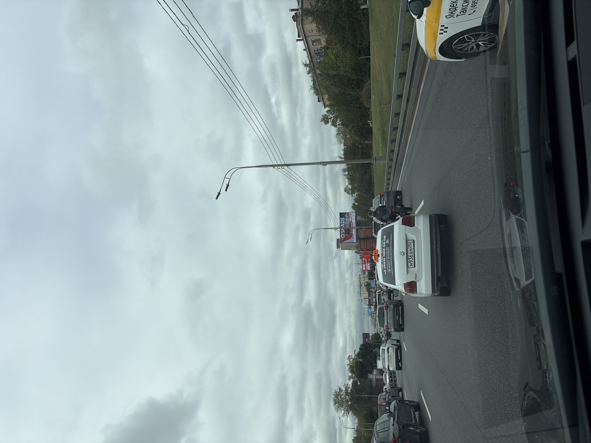 The image captures a traffic jam on a multi-lane road in Moscow, Russia, under a cloudy sky. In the foreground, the windshield of a vehicle shows a reflection of the traffic ahead, including a white Volkswagen Polo taxi with "Яндекс Такси" branding. The taxi has a Russian license plate M936AM250. Numerous other cars are visible in the lanes, all slowed or stopped. On the right side of the road, a raised grassy area separates the road from a building with multiple windows and a red-tiled roof, partially obscured by trees. On the left side of the road, another building and trees are visible, with billboards in the distance. Streetlights with multiple bulbs are present, and power lines stretch across the sky. A person on a bicycle is visible in the left lane. The overall impression is a typical urban scene with heavy traffic.