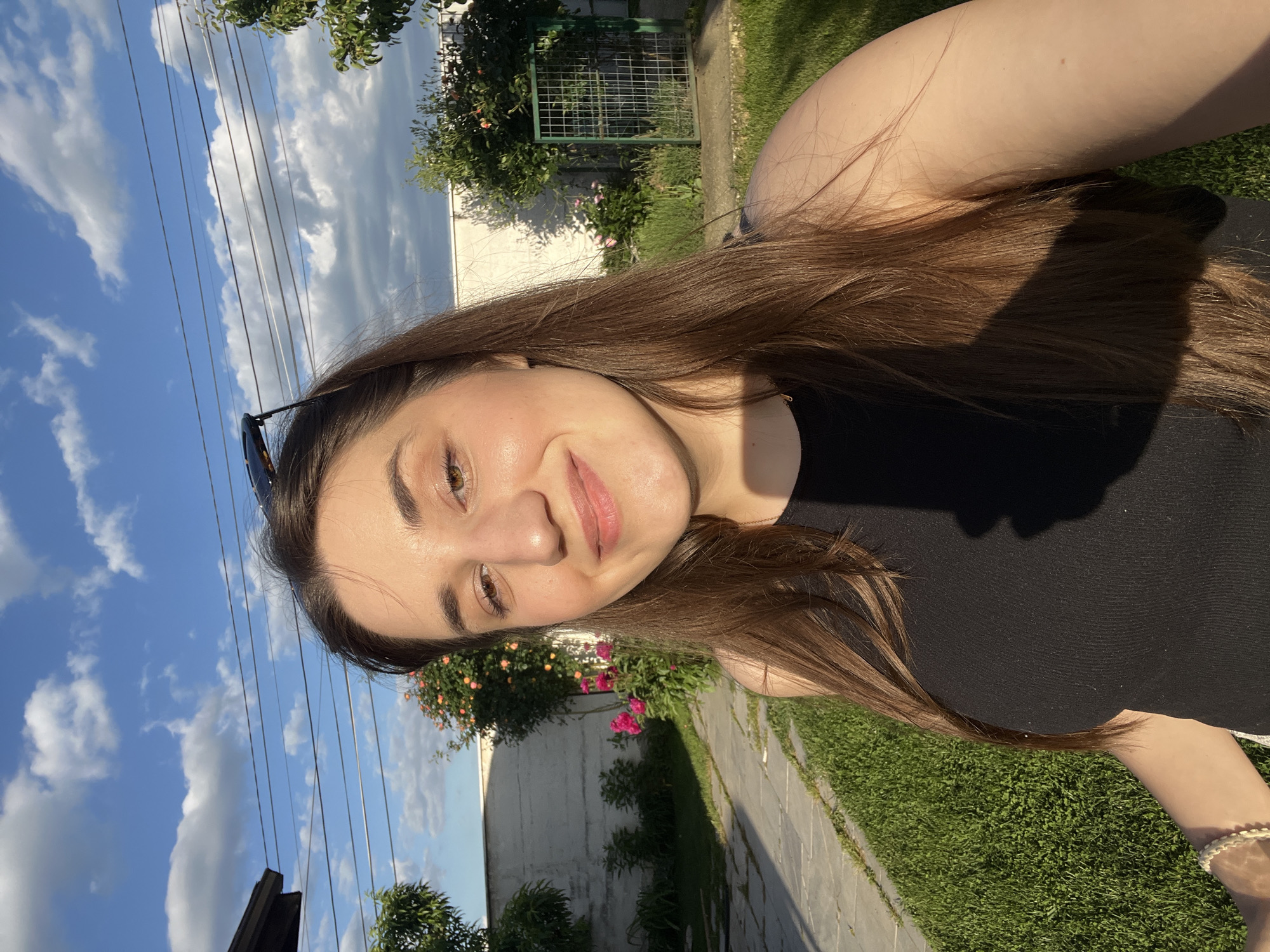 A young woman is the primary subject of the image, appearing to be in a self-portrait or selfie. The background suggests an outdoor environment, potentially in a residential area. The presence of the sky, a fence, greenery, and a sidewalk indicate the setting could be a yard or a park alongside a street. The image captures a sunny day, as evidenced by the shadows and the bright sky. The location is possibly in or around the city of Busmantsi, Bulgaria, as indicated by the prompt. The woman is smiling, which suggests a positive emotion or a deliberate pose for the photo. The overall scene is typical of a casual, everyday setting.
