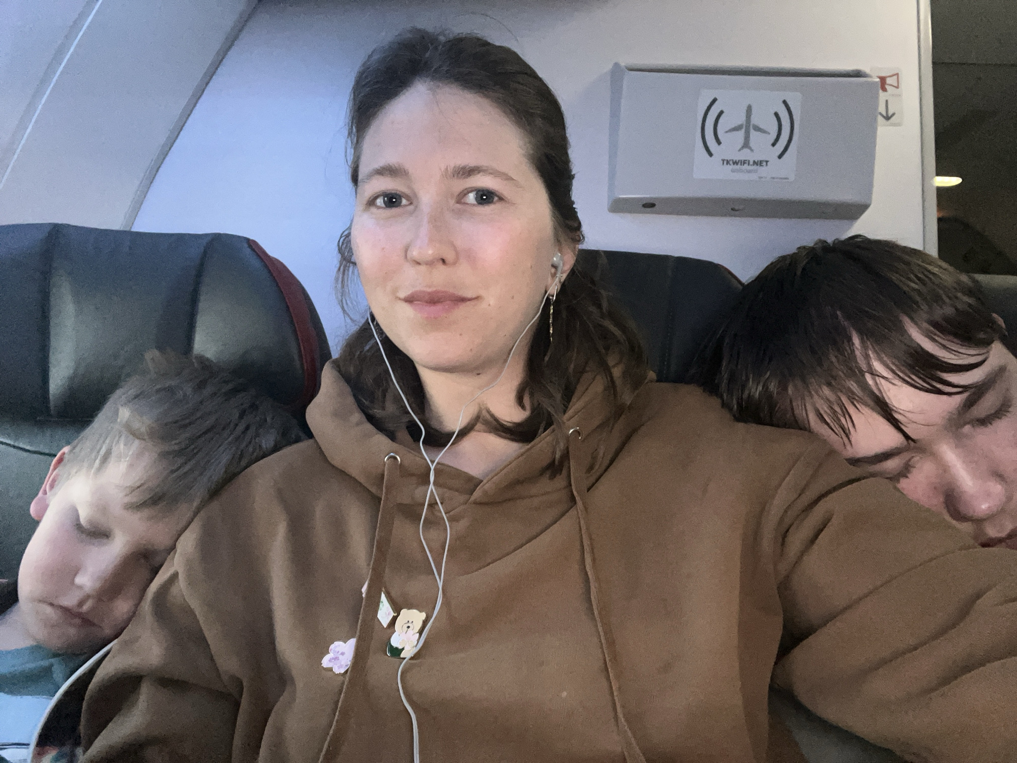The image is a selfie taken inside an airplane cabin, featuring a woman and two sleeping children. The woman, with dark hair tied back, is centered, looking calmly at the camera. She wears a brown hooded sweatshirt with white wired earphones in her ears. Two small pins, one a light pink flower and the other a white teddy bear, are attached to her sweatshirt.
On her left, a child with light brown hair is sleeping peacefully, resting their head on her shoulder. On her right, a second child with dark hair is also asleep, leaning against her, with their face partially obscured. Both children appear to be in deep slumber.
The setting is clearly an airplane, indicated by the typical cabin architecture, including parts of the seats and an overhead compartment. A prominent white sign on the overhead compartment displays a black airplane icon radiating signal waves, with the text "TKWIFI.NET" below it. A small red symbol with a downward arrow is also visible near the top right of the compartment. The cabin lighting is dim, suggesting a nighttime flight or a period of rest. The overall scene depicts a quiet moment during travel, with the woman awake while her young companions sleep.