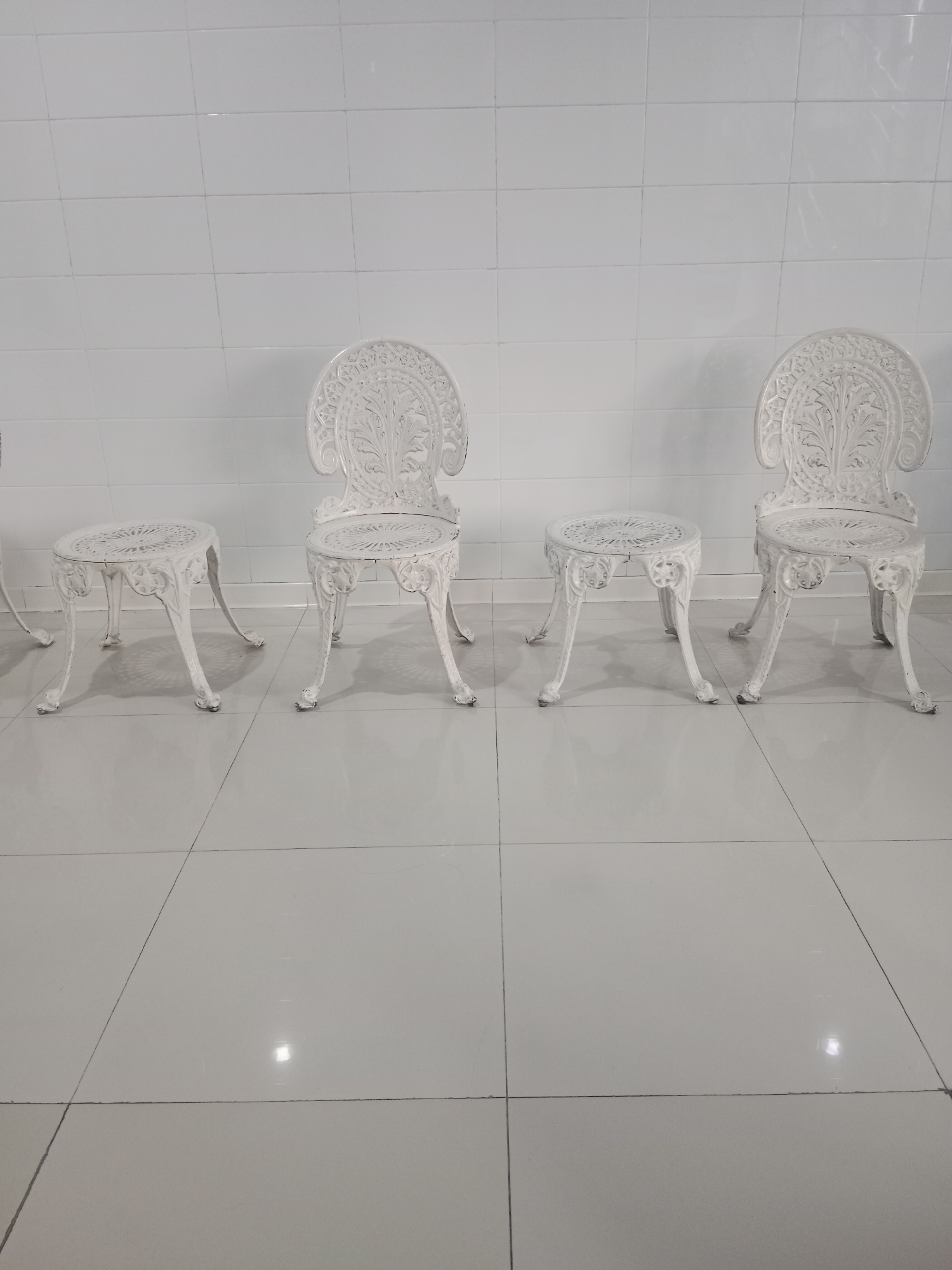 Four ornate white cast iron chairs and three matching tables are arranged on a tiled floor in front of a white tiled wall. The chairs and tables exhibit intricate floral and scrollwork patterns. The scene appears to be indoors or in a covered outdoor area with bright overhead lighting, indicated by the reflections on the floor and wall. The absence of people suggests a staged display or storage. The location is not determinable from the visual information provided.
