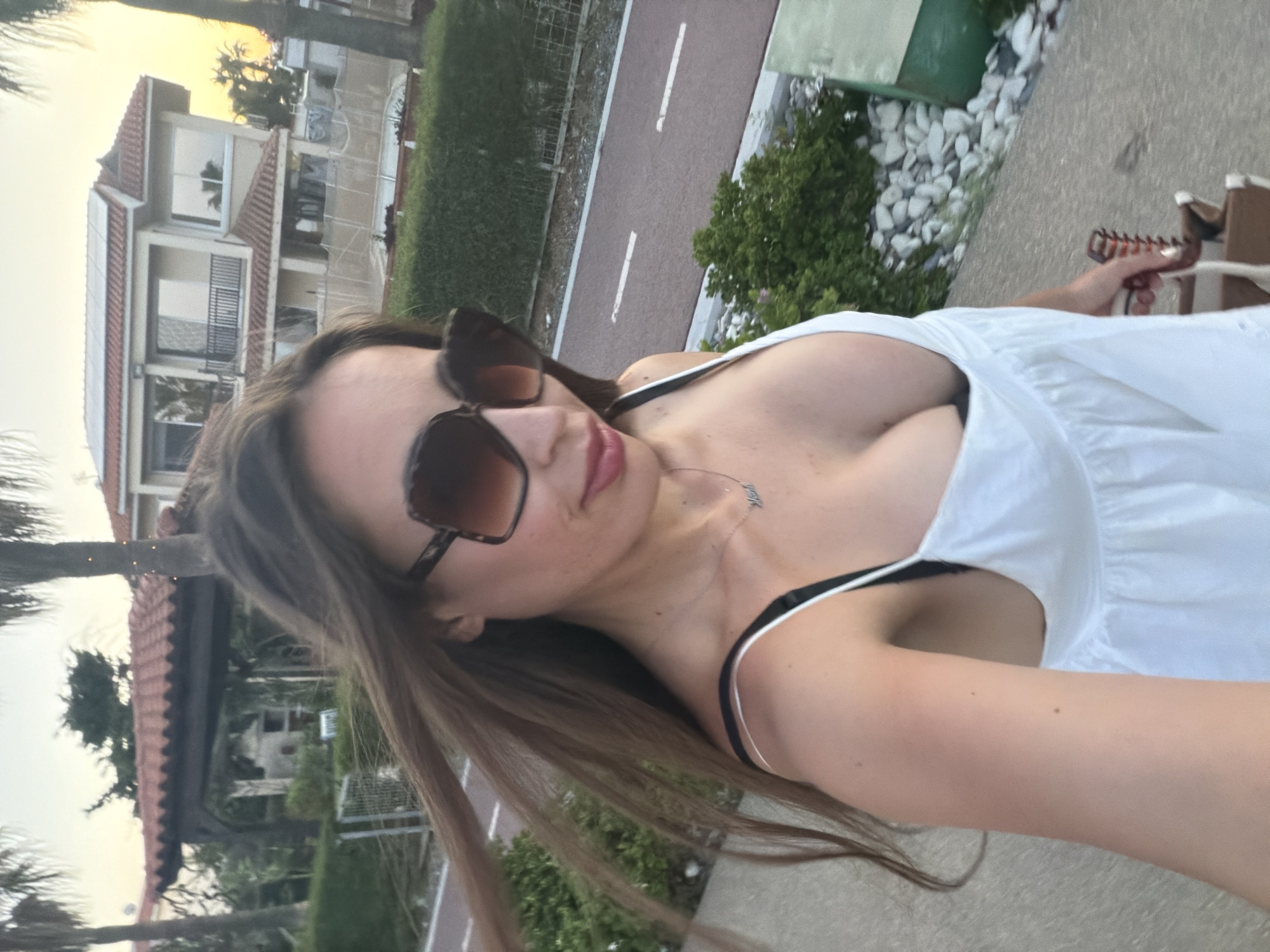 An adult female is captured in a selfie outdoors in Meneou, Cyprus, during sunset or dusk conditions. Her long, brown hair, parted on the side, is wind-blown, with strands visible across her face and to the left. She wears large, square-framed sunglasses with brown-tinted lenses and a tortoise-shell pattern. Her lips are slightly parted, revealing a light pink lipstick and the tip of her tongue. She is wearing a white, loose-fitting top or dress with thin straps, under which a black bra strap is visible on her right shoulder (viewer's left). A silver necklace with a name pendant, appearing to read "Madi" or similar, rests on her chest. Her left hand, with white-painted fingernails, holds a light-colored bag or basket with a dark brown handle, positioned in the lower right corner of the frame.
The background features Mediterranean architecture and landscaping. To the left, a dark-tiled roof and a wooden archway of a building are partially visible amidst green foliage and palm trees. To the right, a two-story, light-colored house with a red-tiled roof and solar panels is prominent. This house includes a balcony and multiple windows reflecting the sky. In front of the house, a construction area with fencing and a partially visible pool or pond can be seen. A reddish-brown paved path, marked with white dashed lines, consistent with a bicycle or pedestrian lane, runs horizontally across the mid-ground, parallel to a grey concrete path where the subject appears to be standing. This path is bordered by green bushes and white decorative rocks. Palm trees are visible throughout the background, silhouetted against a sky that transitions from light blue to warm yellow-orange hues, indicative of sunset or dusk.