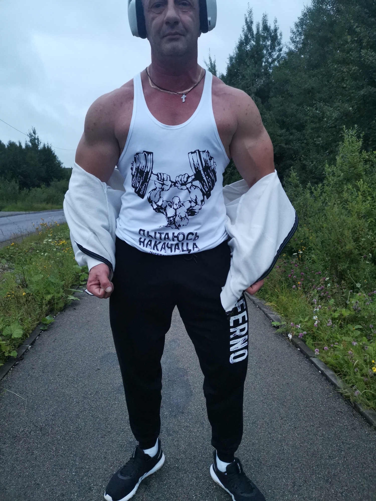 A man is standing on a paved path, which appears to be a sidewalk or a rural road, with a grassy area and trees lining one side. He is wearing a white tank top with a graphic of a barbell and text, black sweatpants with "FERNO" written vertically on the left leg, black athletic shoes with white soles, and headphones. A white zip-up jacket is partially open and draped over his shoulders. The man has a muscular physique and is wearing a silver chain with a cross pendant. The overall lighting suggests it is daytime with overcast skies. The location in Kizel, Russia, is unknown based on the provided image.