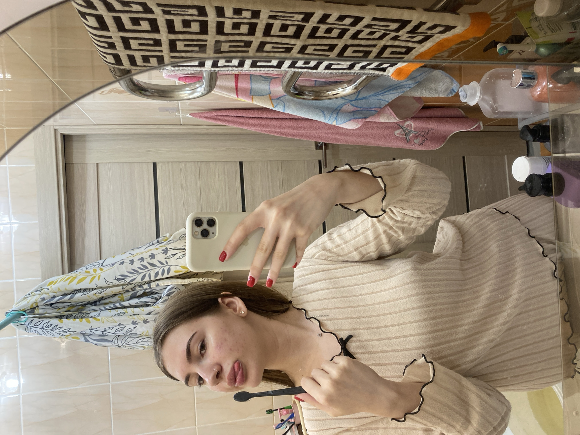 The image captures a young woman taking a mirror selfie in a bathroom in the city of Unknown, Unknown. The woman, appearing to be in her late teens or early twenties, has long brown hair, a silver nose ring on her left nostril, and a small gold stud earring in her right ear. She is wearing a light beige, vertically ribbed long-sleeved top with black ruffled trim along the collar and cuffs. Her fingernails are painted red. With her left hand, she holds a light-colored smartphone to capture her reflection. In her right hand, she holds a black toothbrush, with her tongue playfully extended.
The bathroom's walls are covered with light beige ceramic tiles. A light wood-grained door with two horizontal panels is visible in the reflection to the left. Hanging above the door is a white towel patterned with grey and yellow leaves. To the right, a chrome towel rail supports multiple towels: a beige towel with a dark brown geometric pattern, a pink towel displaying a graphic, a light blue towel with a printed design, and a partially visible orange towel. Various hygiene products, including several pump bottles and containers, are arranged on a clear glass shelf or countertop beneath the mirror.