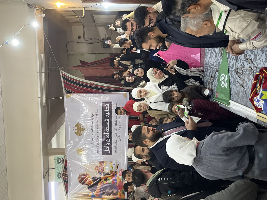 An indoor scene in Kafr Batna, Syria, depicts a crowded community event. The room has light-colored walls, fluorescent lighting, and string lights overhead. A large white banner on the back wall features Arabic text, including "برعاية محافظة ريف دمشق" (Under the patronage of Damascus Countryside Governorate) and "فعالية فسحة أمان وأمل" (Activity: Space for Safety and Hope), alongside a graphic of a child playing with blocks and a golden eagle emblem (Syrian coat of arms). Flanking the banner are patterned fabric drapes or tent-like structures.
In the immediate foreground, a man in a dark suit with a beard is handing a small white and green toy airplane to a woman wearing a grey coat and a white hijab. A young child with faint face paint resembling whiskers stands beside them, observing the interaction. To the right, another man in a dark jacket is shaking hands with an older man dressed in a khaki shirt with a green scarf and badges, typical of a scout uniform.
A table covered with a patterned cloth runs along the foreground, displaying various objects including small green flags with a white star, crescent, and ribbon emblem, and multiple small metal items resembling clasps or hooks. Several scout-style neckerchiefs and badges are also visible on the table.
The background is densely populated with men, women, and children. Many women are wearing hijabs. Individuals are observing the activities, conversing, and some are holding up smartphones to record or photograph the event. The overall atmosphere appears organized and engaged.