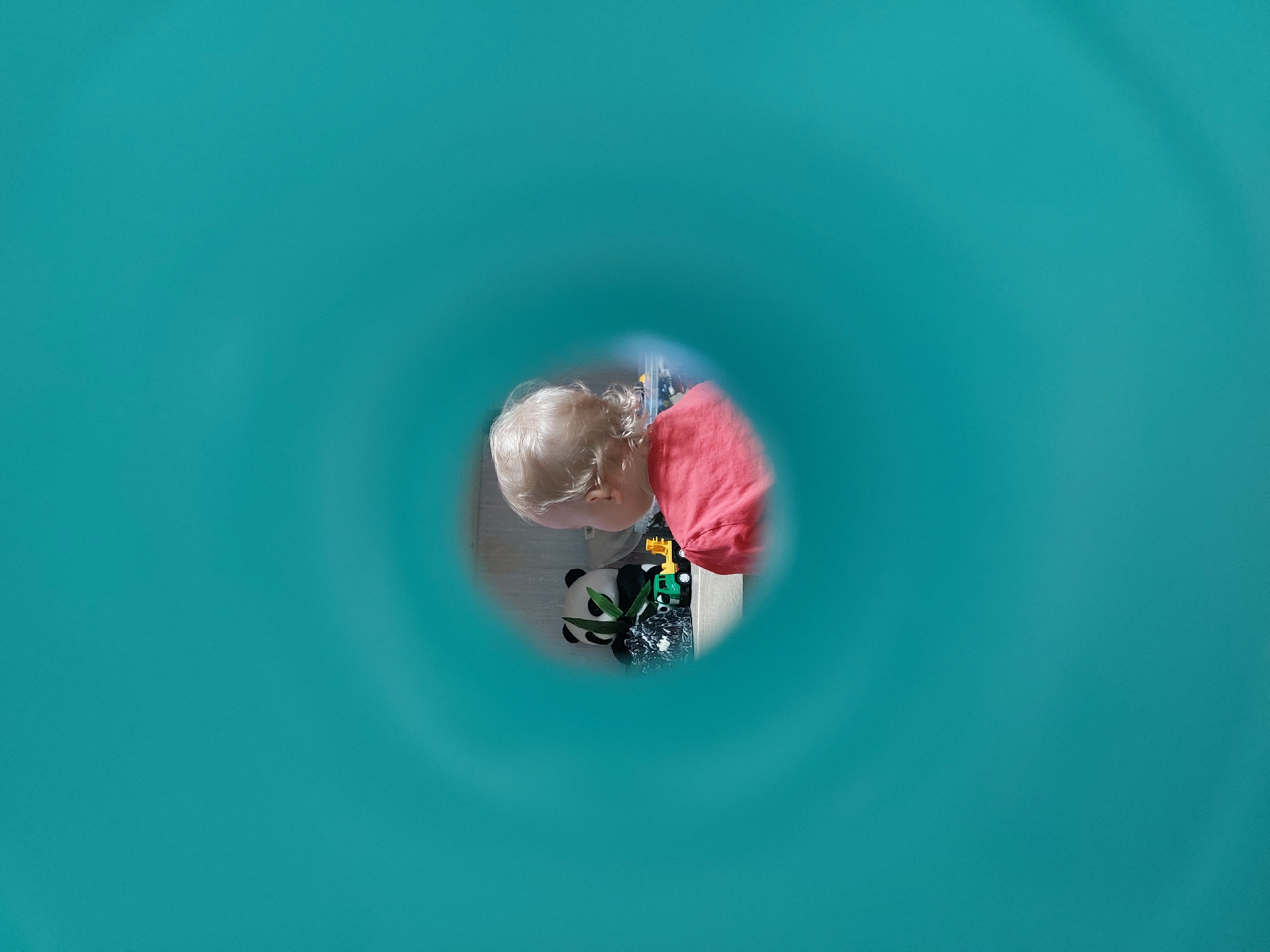 A child with blonde hair is looking through a circular hole in a turquoise-colored surface. The child is wearing a red shirt and is looking down at a panda stuffed animal and a yellow toy truck. The location is Chaikovsky, Russia.