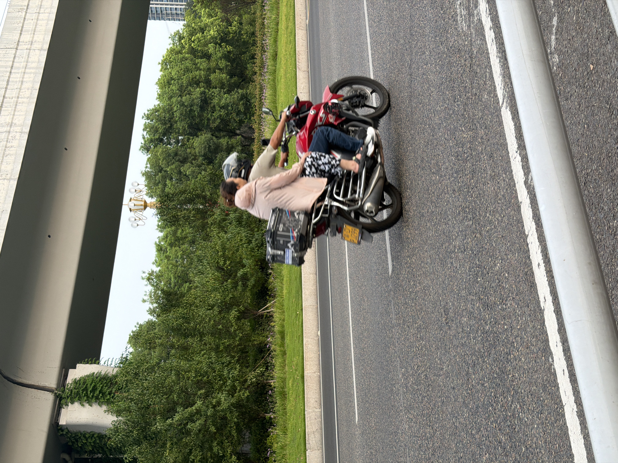 Two individuals, a man and a woman, are riding on a red motorcycle on a paved road in Jiangjin, China. The man is driving, wearing a light-colored t-shirt and blue jeans, and a helmet. The woman is seated behind him, wearing a light pink jacket and patterned pants, with her hair tied up. She is wearing sandals. The motorcycle has a black storage box mounted on the back and a yellow license plate. The road has white lane markings. In the background, there is a grassy embankment with trees and a large overpass structure. A building is visible in the far distance to the right. The scene appears to be during the daytime, under an overcast sky.
